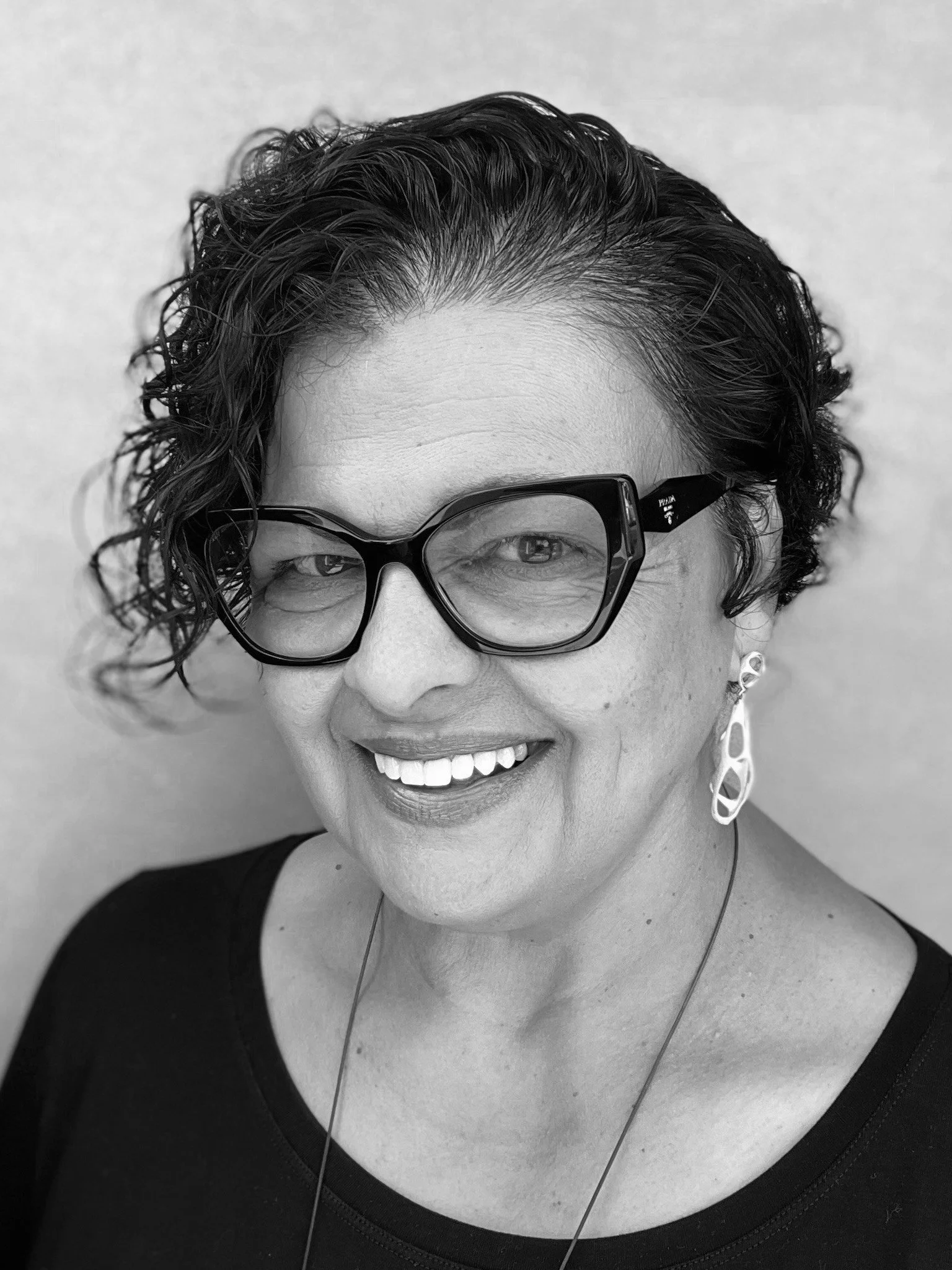 A black-and-white portrait of a woman with short curly hair wearing large glasses, earrings, and a black top, smiling at the camera.