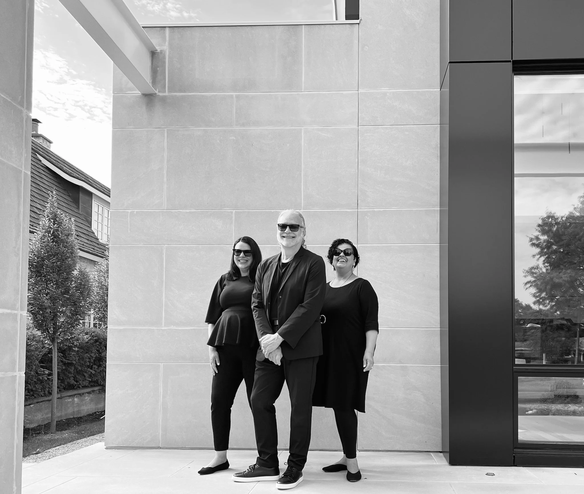 Three people standing outdoors in front of a modern building: an older man in the center, a woman to his left, and a woman to his right. They are all wearing black clothing and sunglasses, smiling at the camera.
