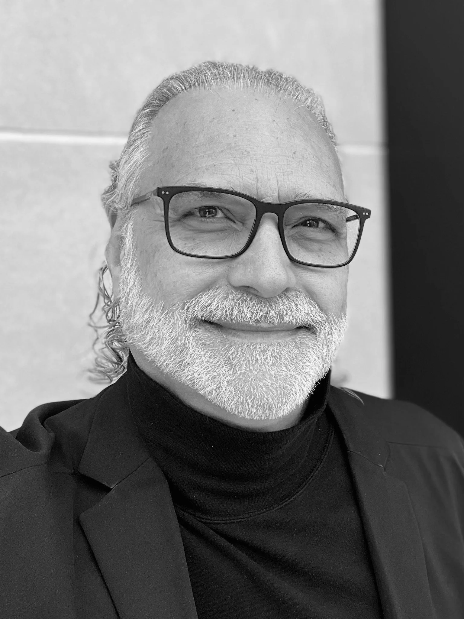 Black and white close-up photograph of a smiling man with glasses, a beard, and long hair pulled back, wearing a black turtleneck and blazer, standing against a light-colored wall.