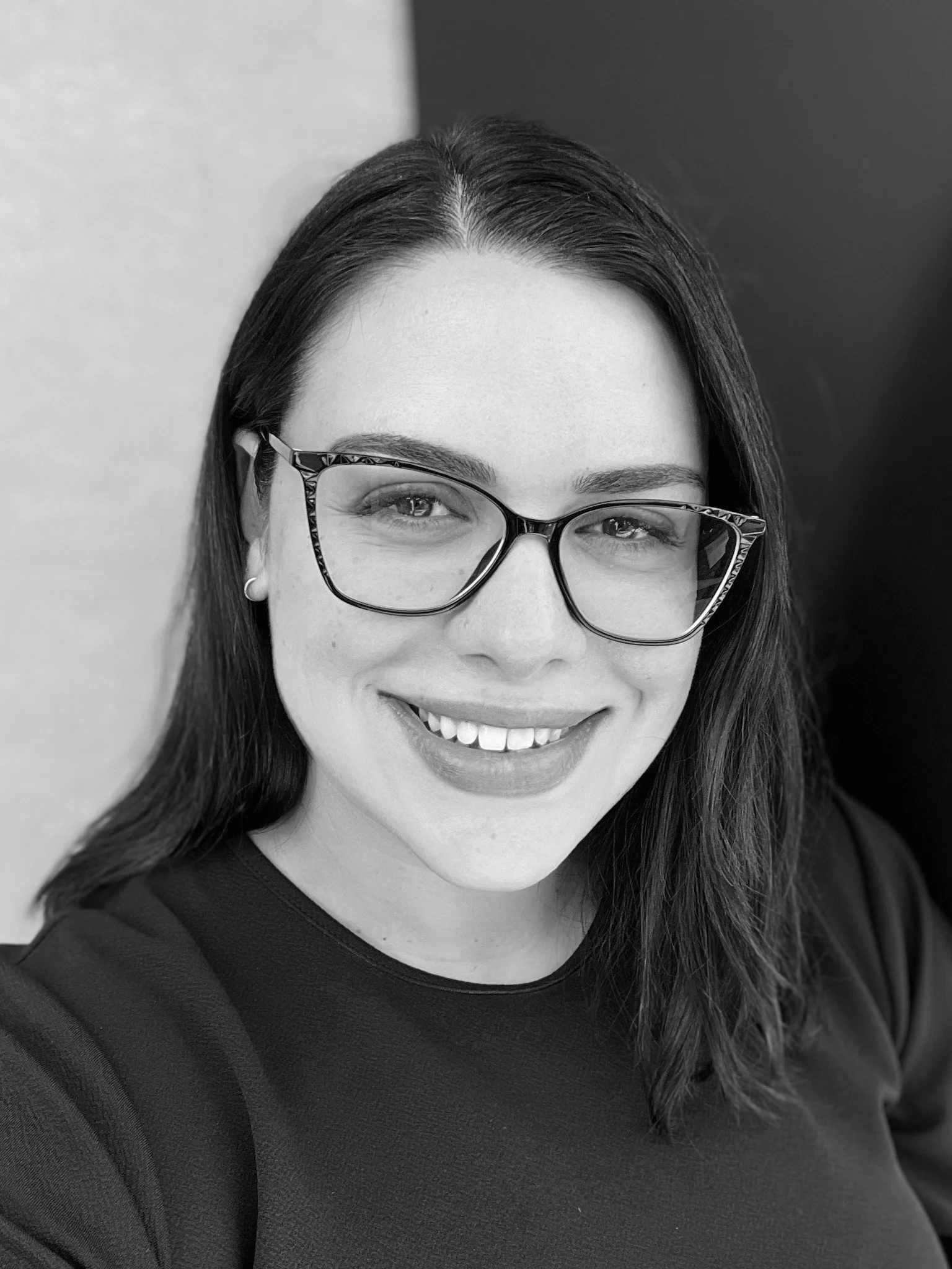 A smiling woman with glasses and long dark hair, wearing a dark top, in black and white.