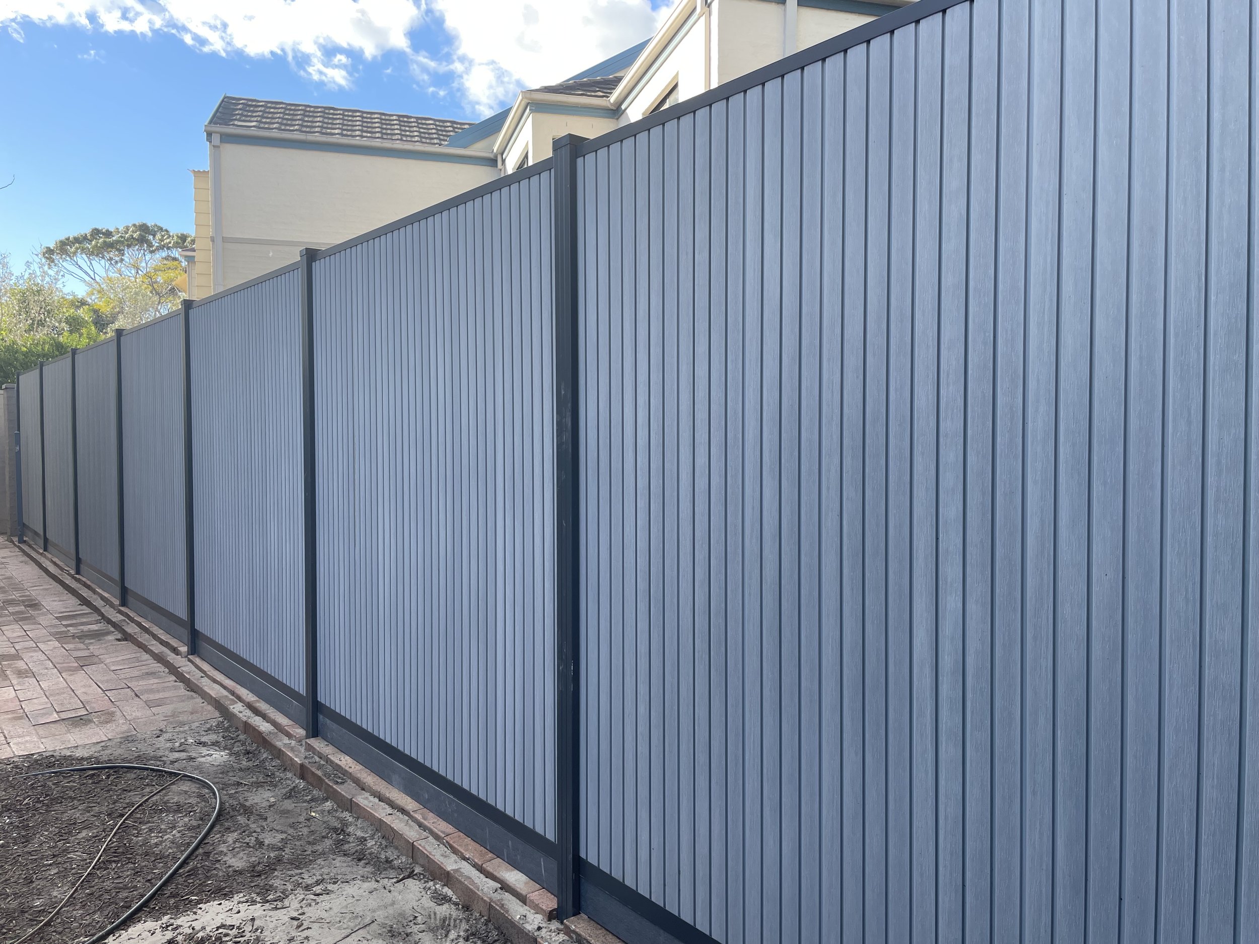 A tall blue privacy wpc fence running along the sidewalk in front of a light-colored house with a gray roof. There are some trees and a partly cloudy sky in the background. The ground in front of the fence shows some dirt and cobblestone paving.