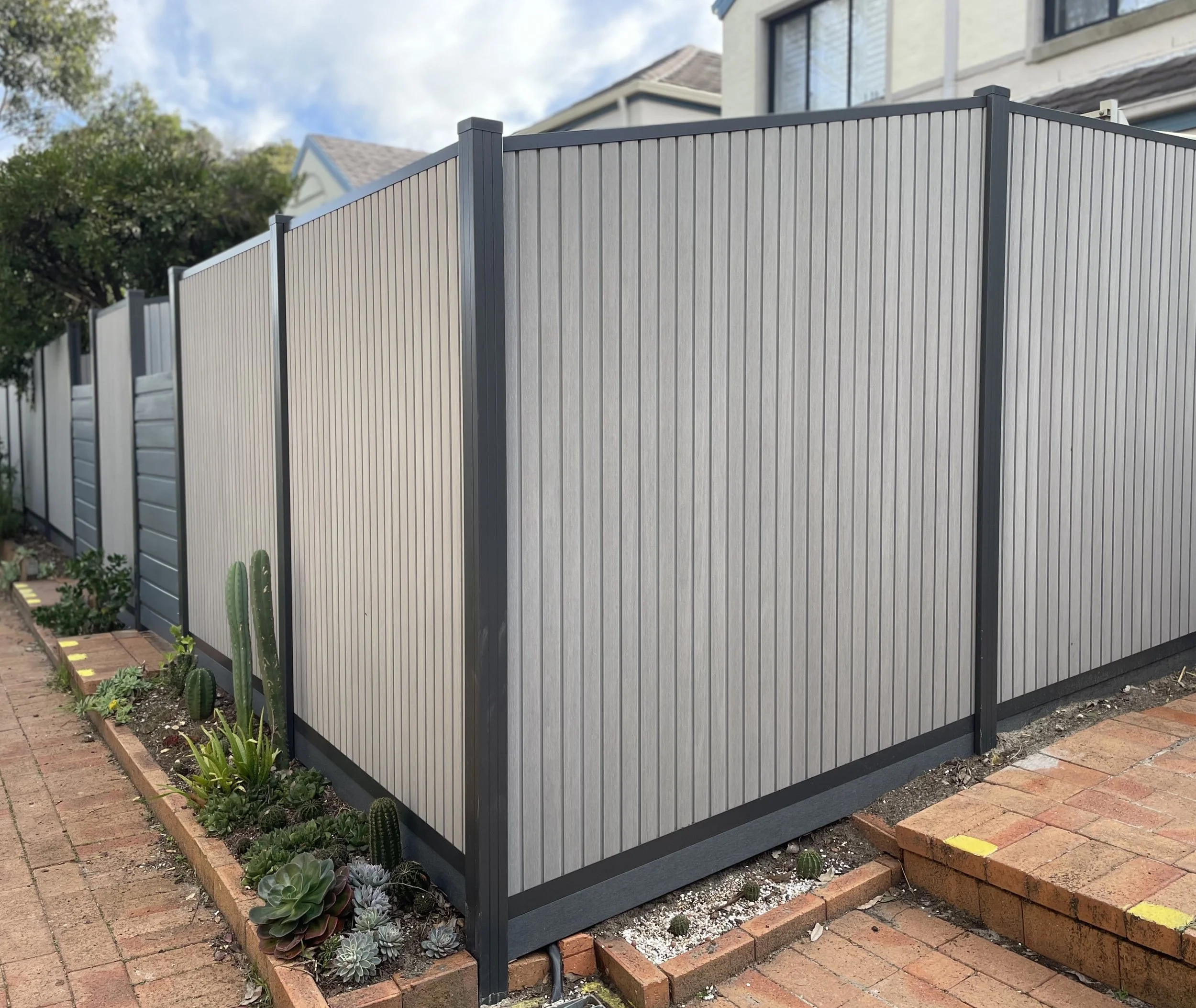 A gray and black wpc privacy fence around a backyard garden, with succulent plants and cacti at its base, in front of a house with a light-colored exterior and large windows.