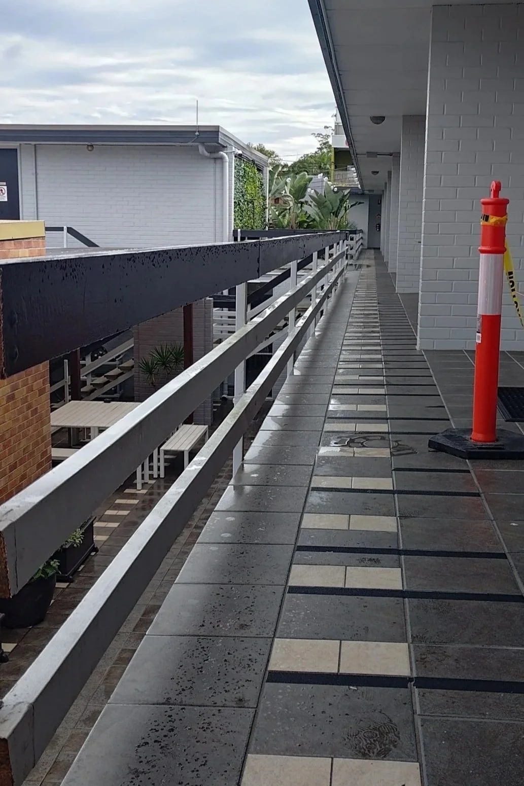 A wet outdoor balcony with tiled flooring, white brick walls, a black railing, and an orange safety pole.