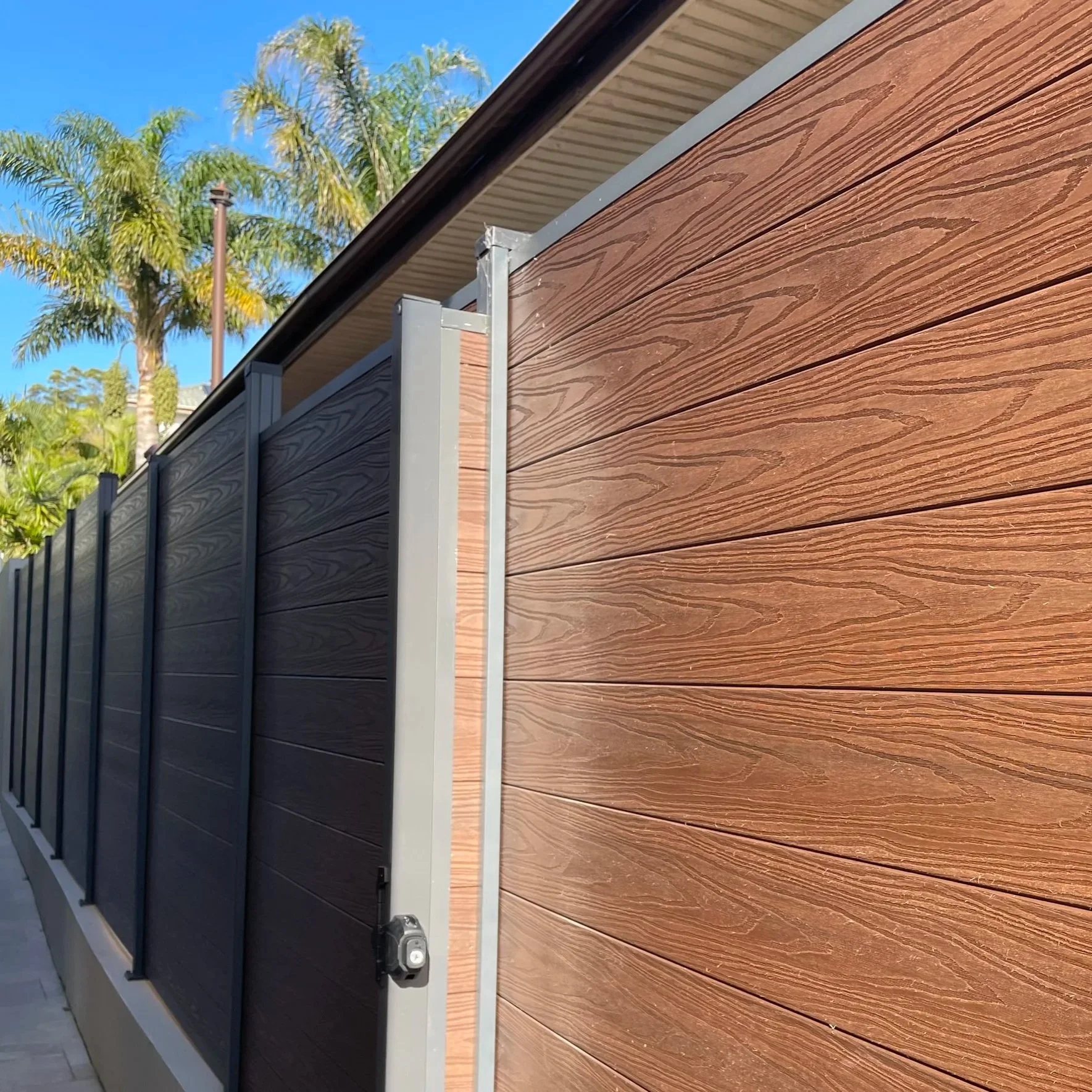 Close-up of a modern wood-plastic composite fence with brown and black horizontal panels, vertical metal supports, and a power socket underneath, with palm trees and a clear blue sky in the background.