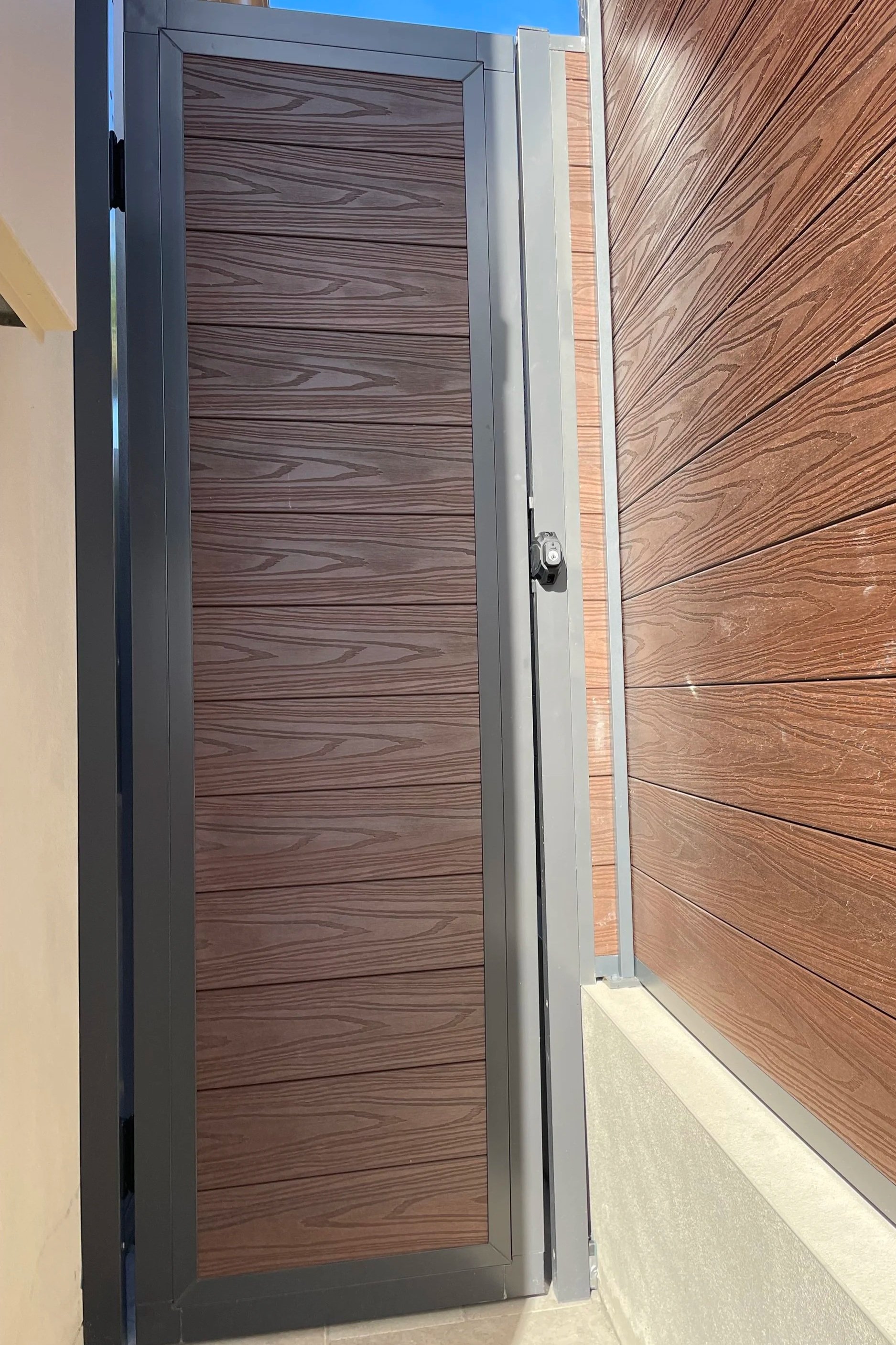 A wpc fence with horizontal grain patterns, a aluminium door frame, a locked security door with a small keyhole, and a wooden floor with planks.