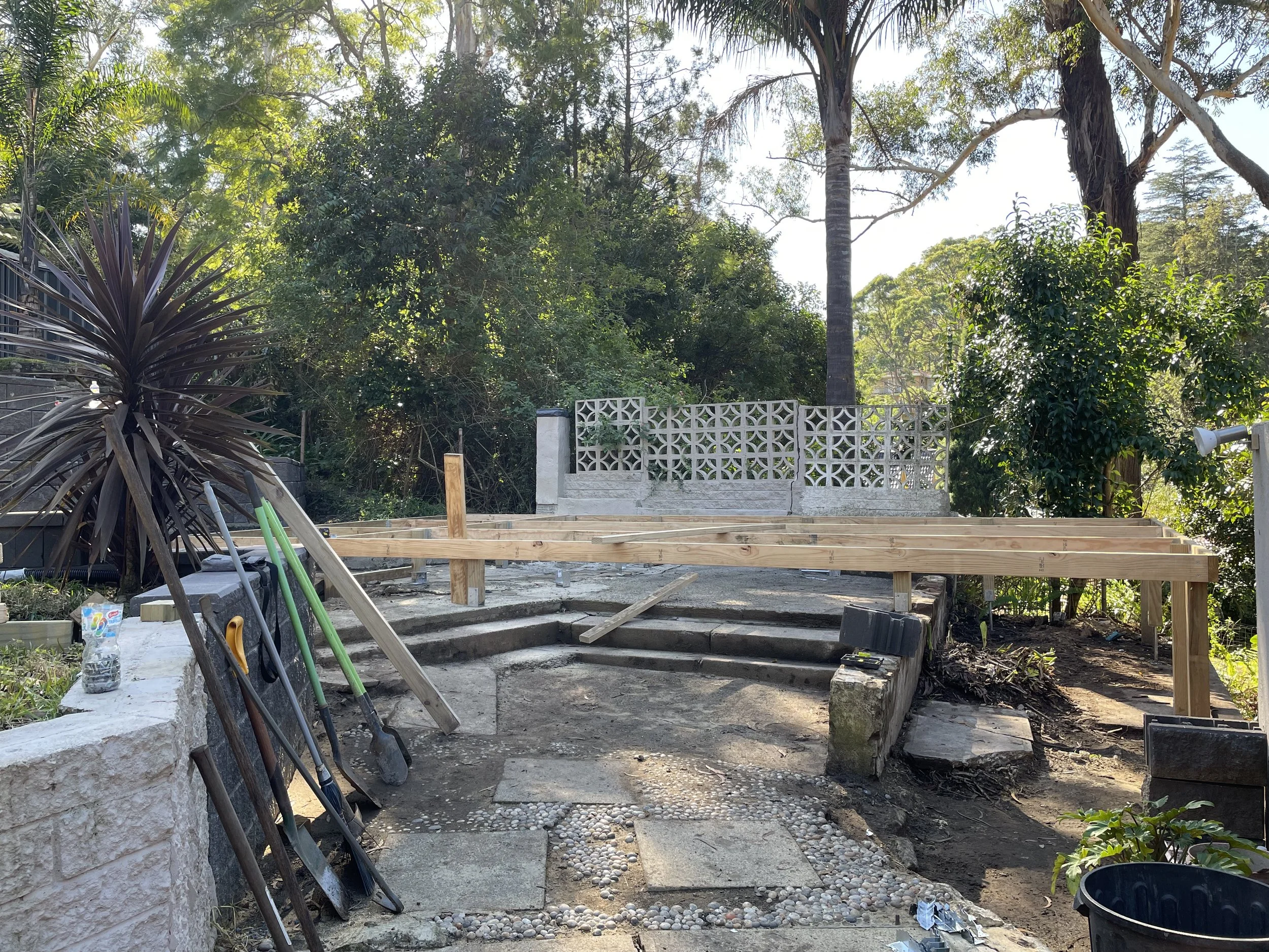 Construction site with wooden framework for a deck, various tools and gardening items in the foreground, and a decorative white concrete block wall with trees and bushes in the background.