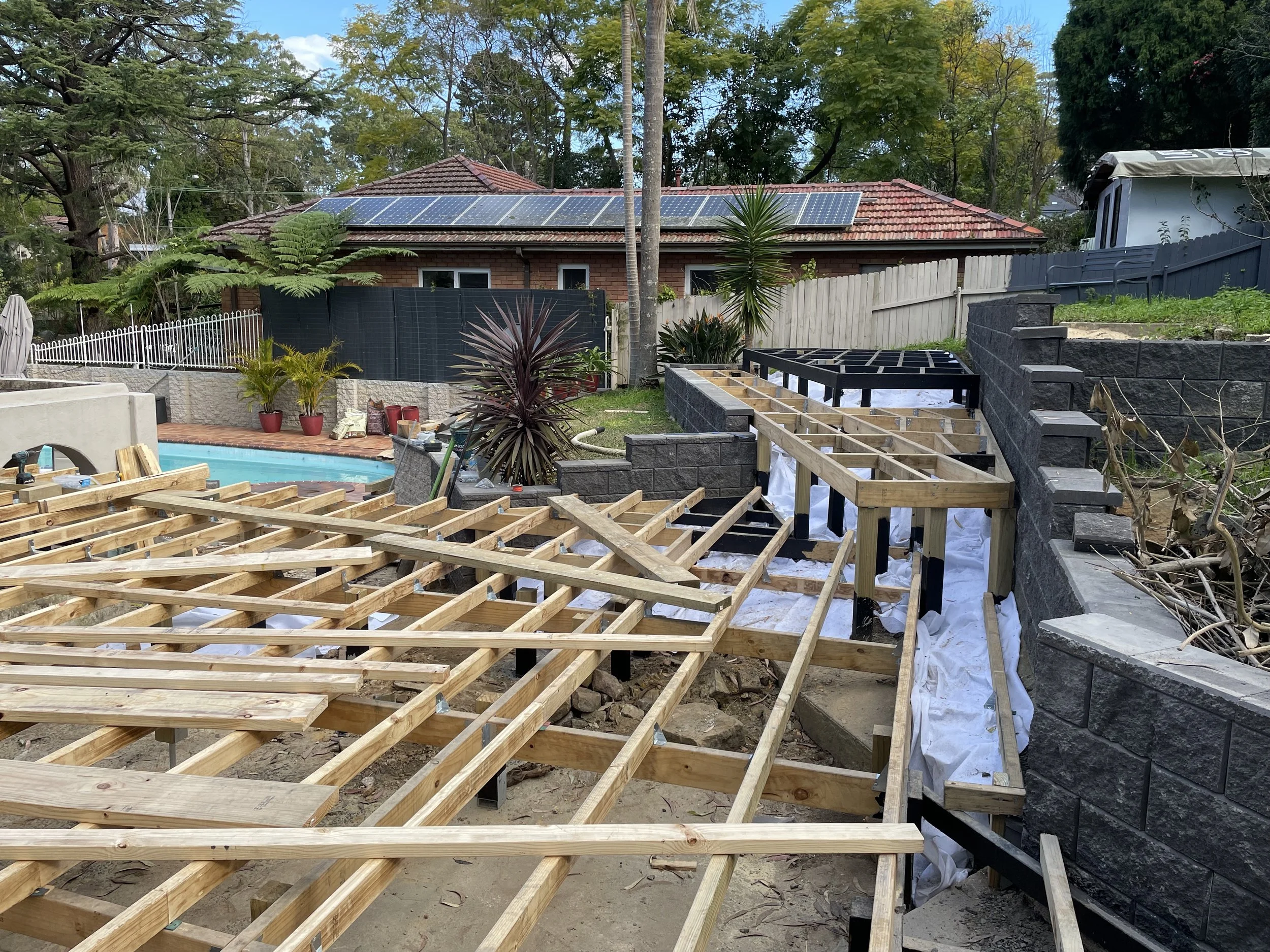 A backyard under construction with wooden framing for a deck or patio, landscaping plants, a swimming pool, and a house with solar panels on the roof.