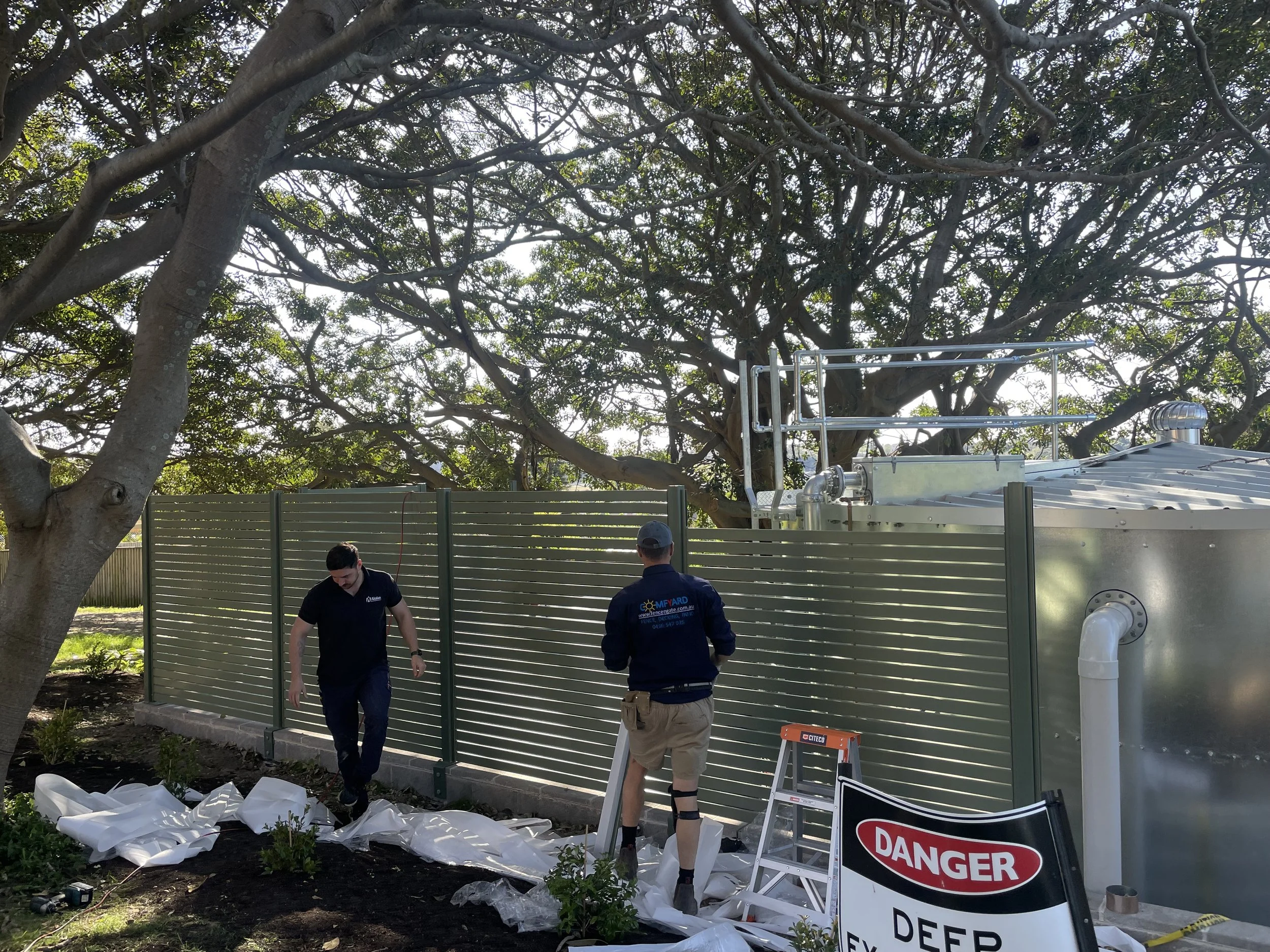 Three workers installing aluminium screens next to a large tree, with equipment and warning signs nearby.