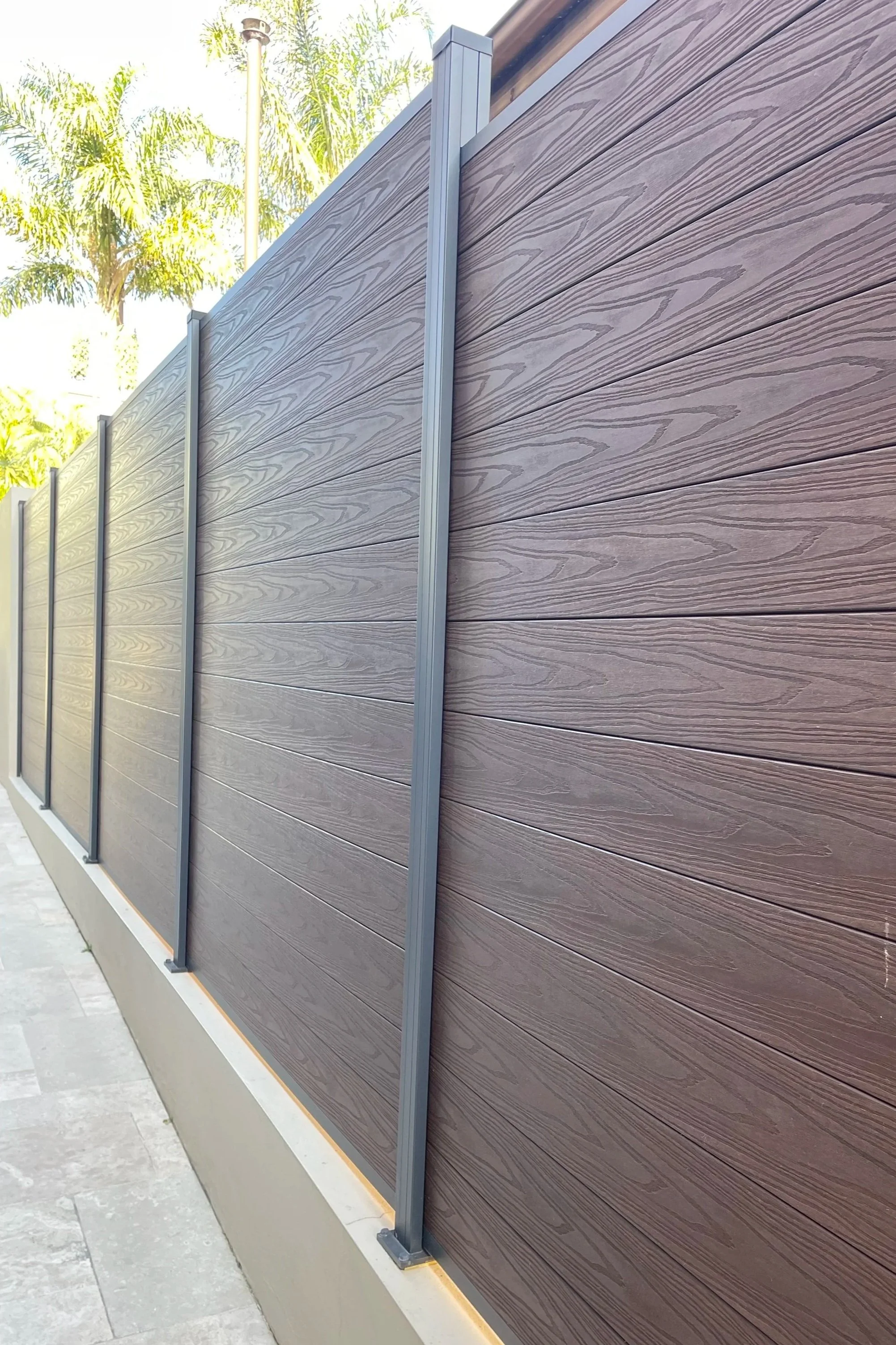 A modern outdoor privacy wpc fence with horizontal wpc panels in a grayish-brown color, supported by aluminium posts and a base, with palm trees and a clear sky in the background.