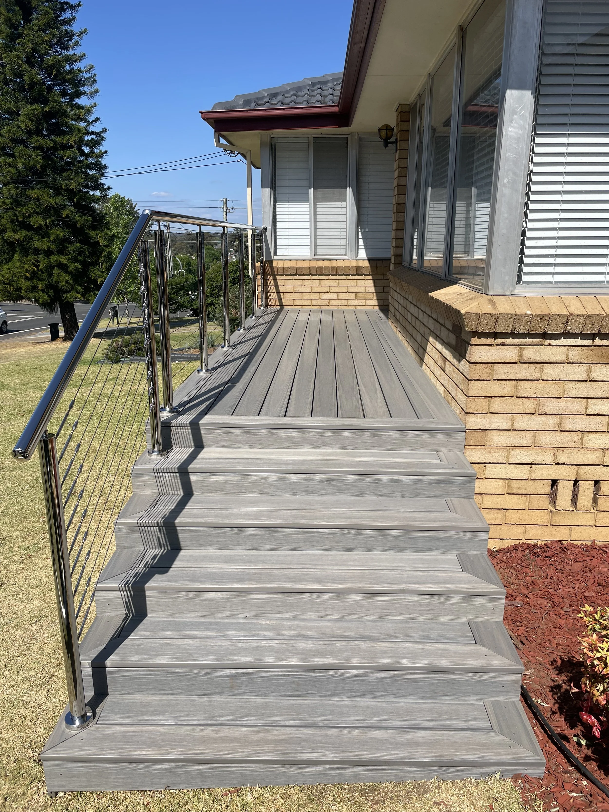 Outdoor grey wooden stairs leading up to a patio with metal railing, attached to a brick house with large windows, on a sunny day with clear blue sky.