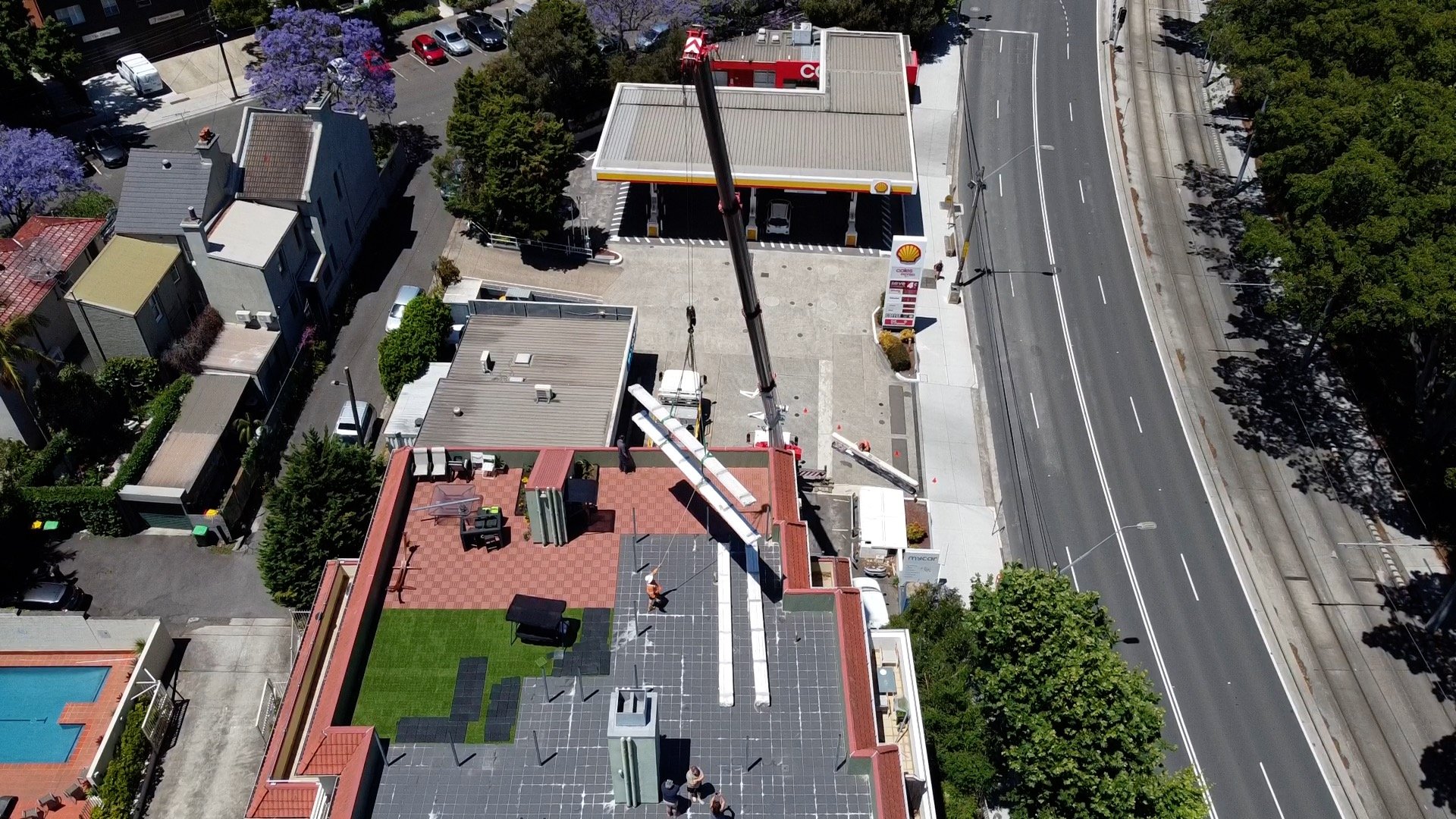 Aerial view of a Shell gas station nearby multi-story building with a crane lifting aluminium panels and posts on the rooftop of this building, adjacent to a sidewalk and busy street with trees.