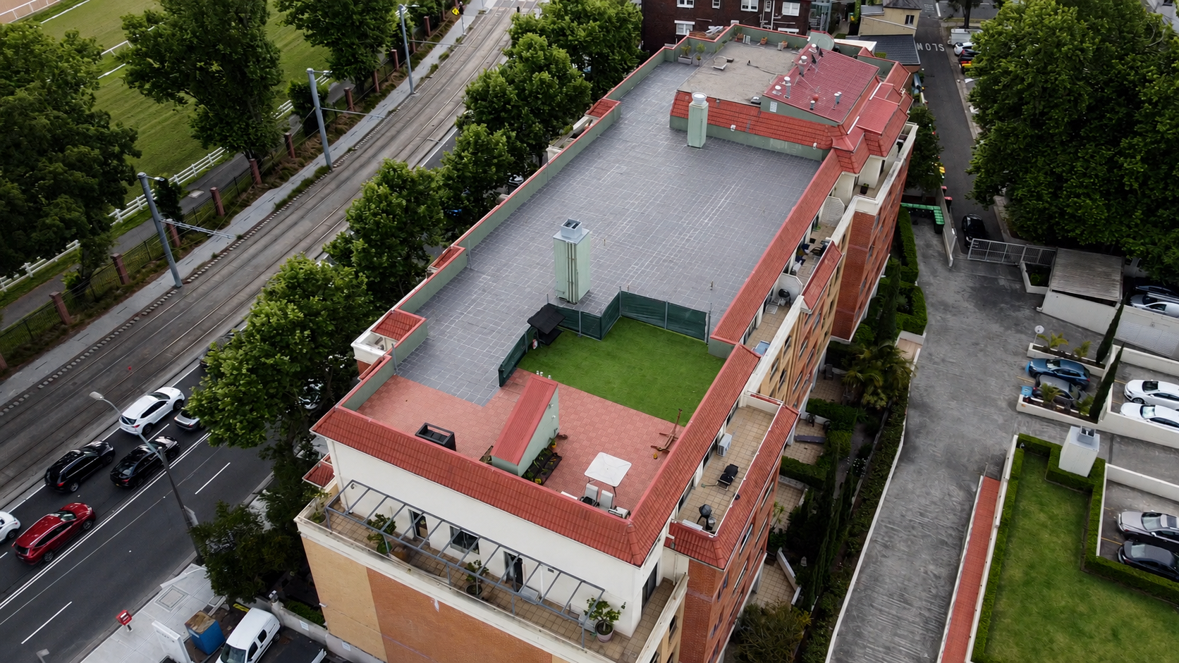 Aerial view of a multi-story building with a large flat rooftop, surrounded by trees, parked cars, and adjacent streets. The rooftop includes a small grassy area, an outdoor seating space with a white umbrella, and various HVAC units.