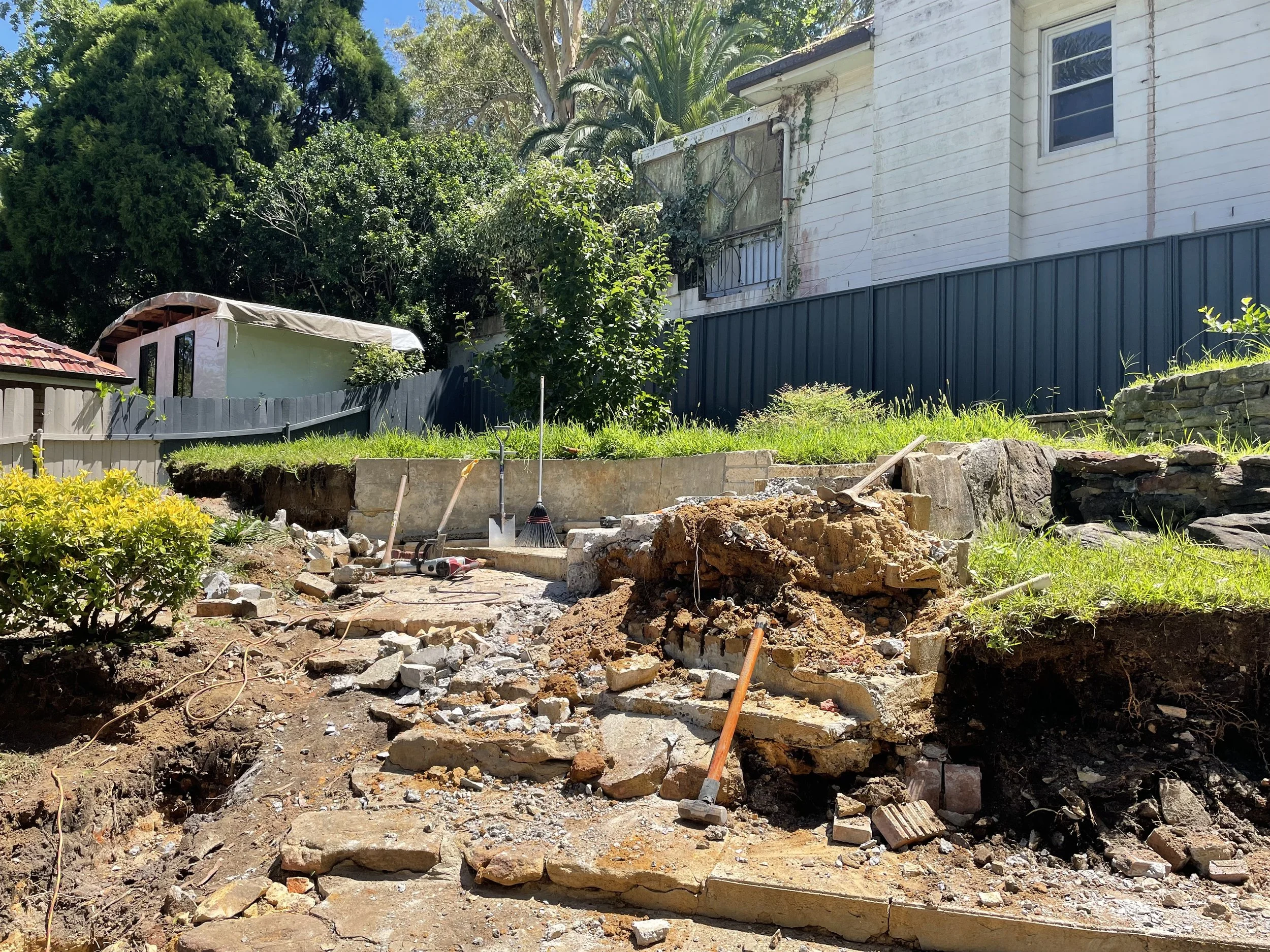 A backyard garden under construction with exposed soil, broken concrete, and construction tools such as a broom and a hammer. There are trees, shrubs, and a white house with a small balcony in the background.