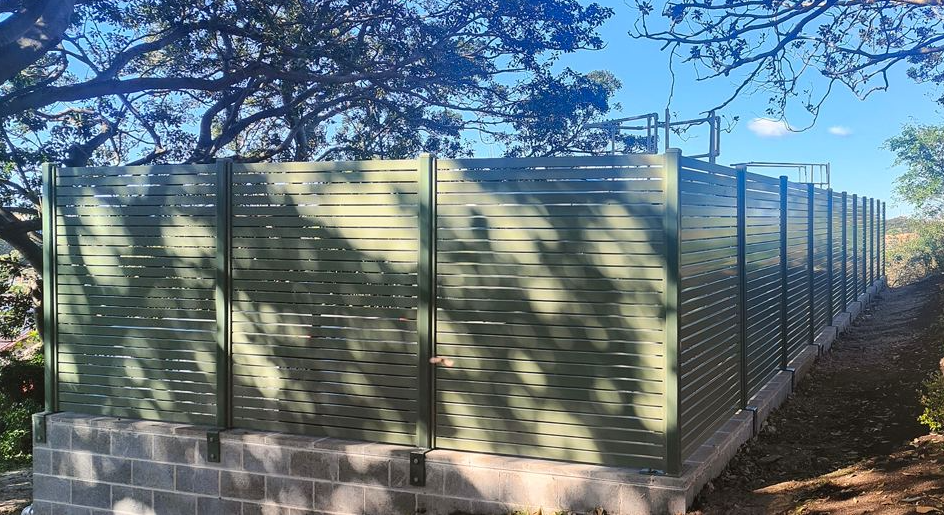 Metal fence with horizontal slats, mounted on concrete blocks, with shadows of tree branches and leaves over it, along a dirt pathway outdoors under a blue sky.