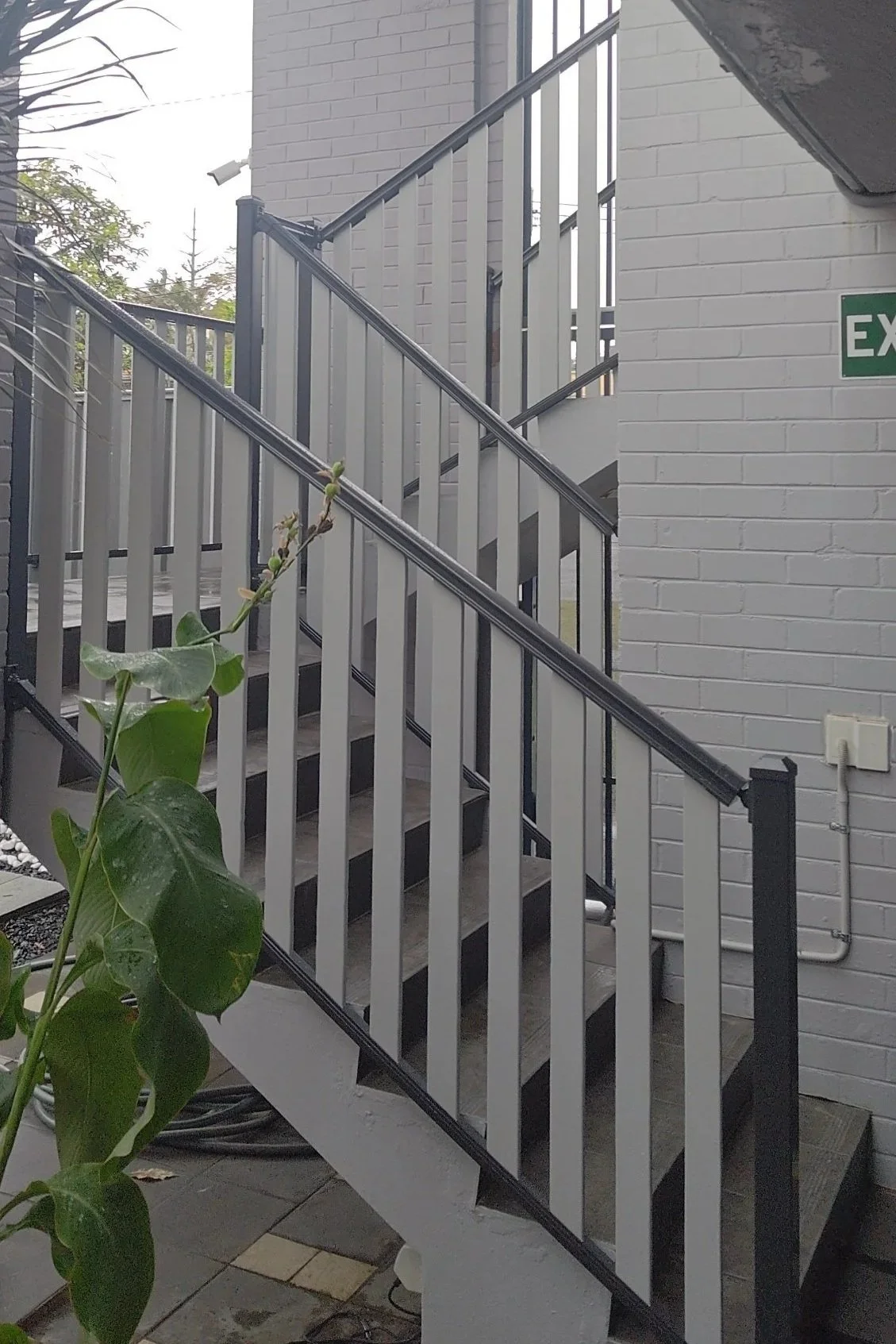 Exterior staircase made with black and white wpc railings, adjacent to a white brick wall with an exit sign.