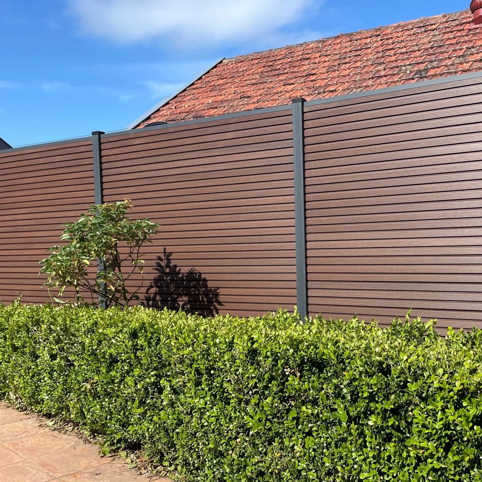 A tall brown privacy wpc fence with horizontal slats, a red-tiled roof, a small green shrub, and a green hedge along a sidewalk, under a blue sky.