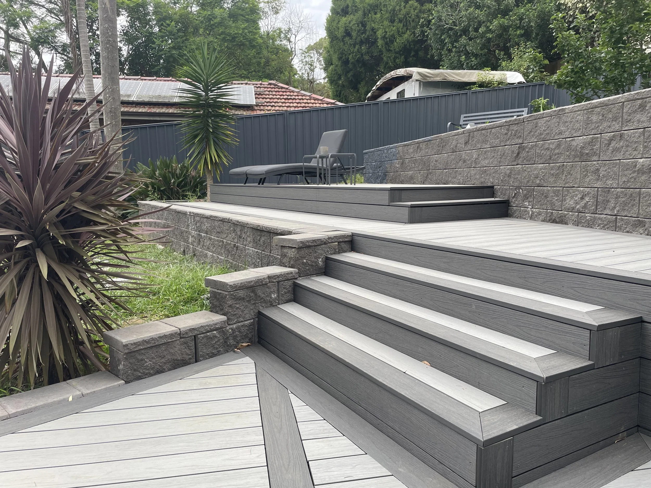 A backyard patio with wooden-plastic composite stairs leading up to a seating area, surrounded by trees and greenery, with a colorbond fence in the background.