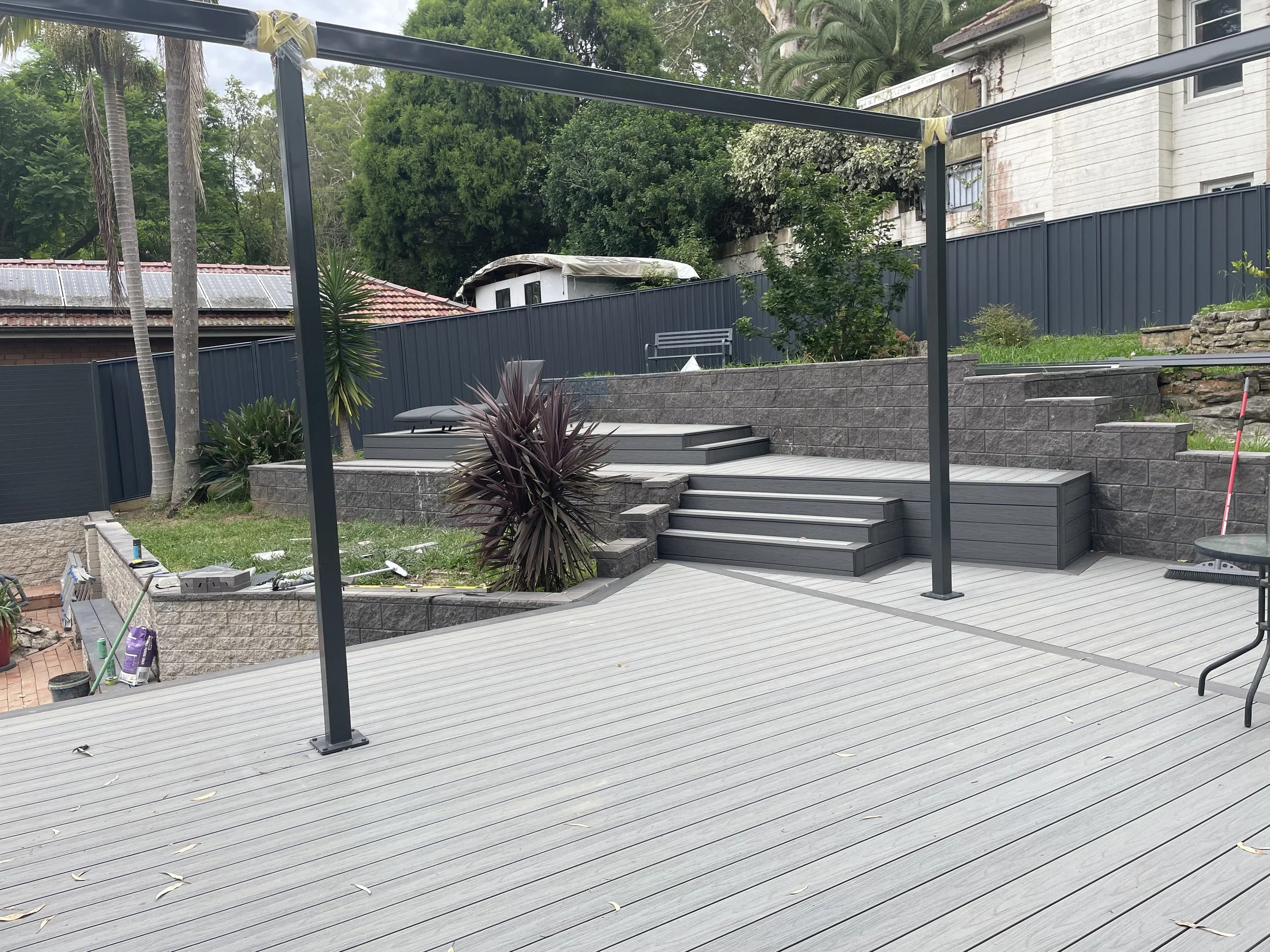 A backyard patio with incomplete structure, metal beams, potted plants, and surrounding greenery.