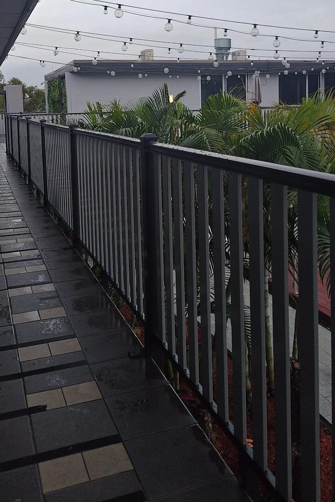 A balcony balustrade with black wpc railing, wet tiled floor, green plants underneath, and string lights hanging overhead on a cloudy day.