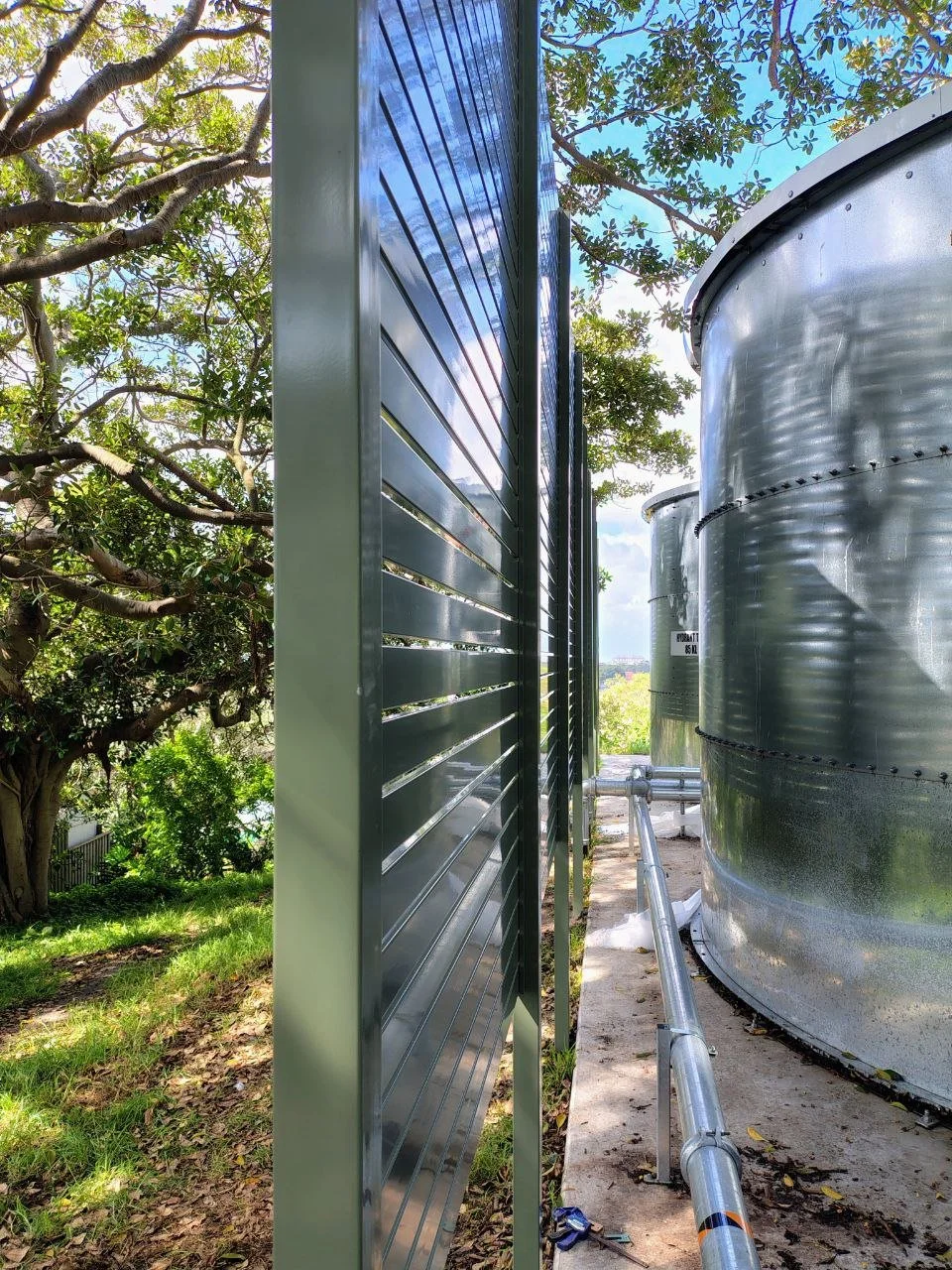 Aluminium screen panels near large cylindrical tanks outdoors on a concrete pad, with trees and blue sky in the background.