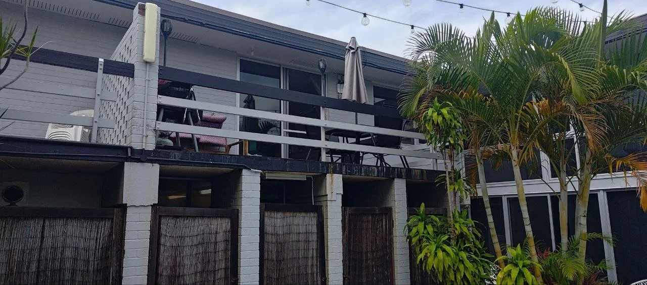 Exterior view of a hotel with a second-floor balcony, patio furniture, and tropical plants in front.