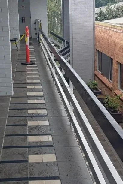A balcony with an old black wood railing next to a brick building, rain-damaged tiled flooring, and a temporary orange safety cone.