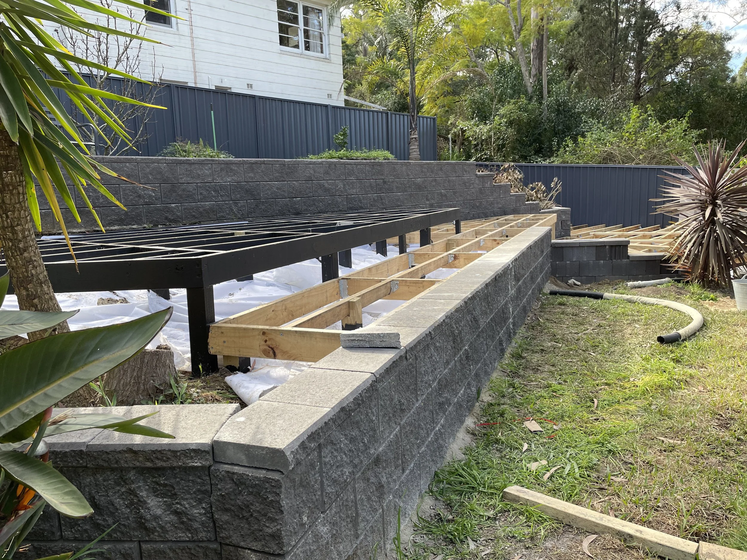 Construction site with a stone wall and wooden framework for decking, surrounded by green plants and trees.