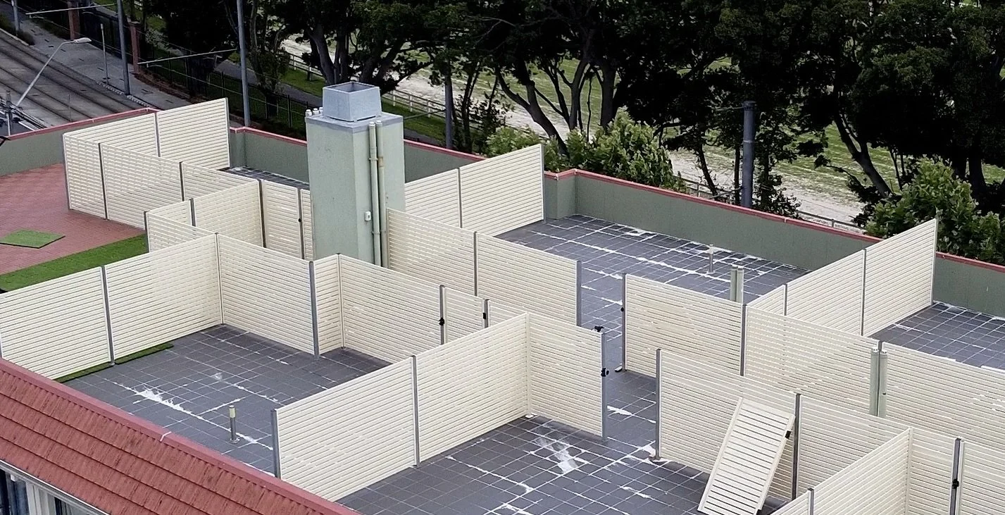 Empty rooftop area with cream-colored privacy screens, a tall vent, and rooftop tiles, surrounded by green trees and a red-roofed building.