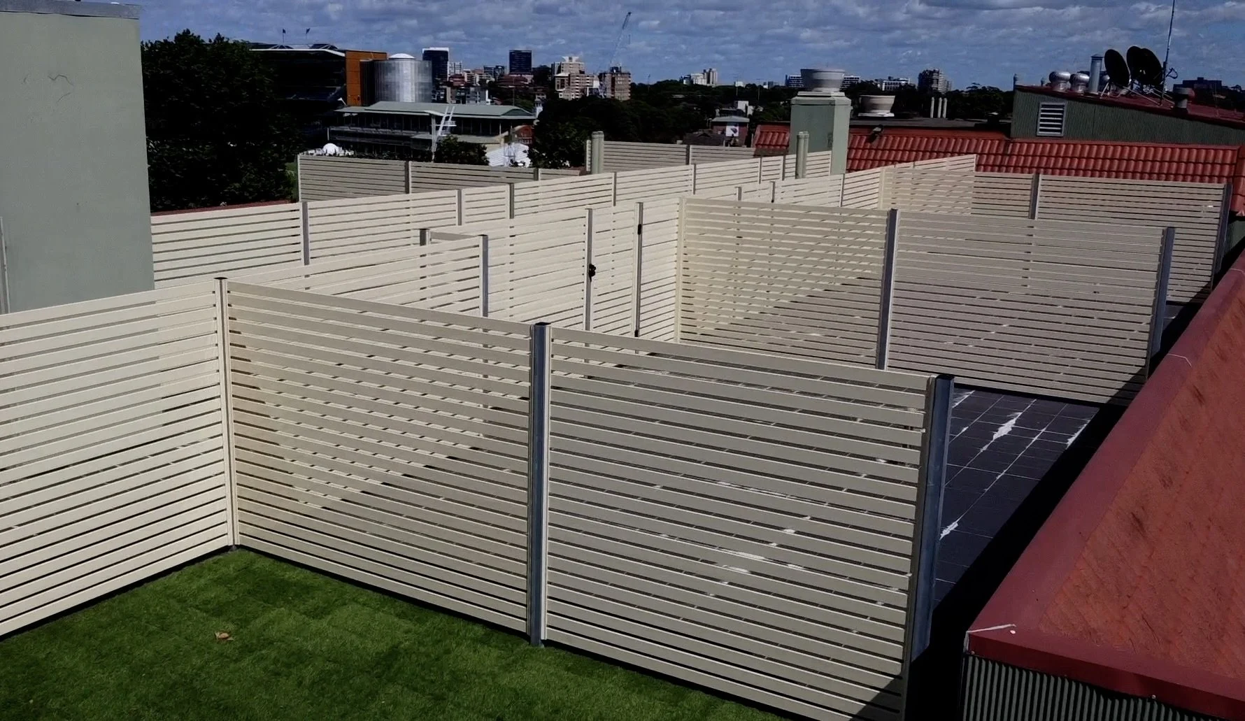 View of a rooftop area with cream-colored privacy fences, a small patch of green grass, and city buildings in the background under partly cloudy skies.
