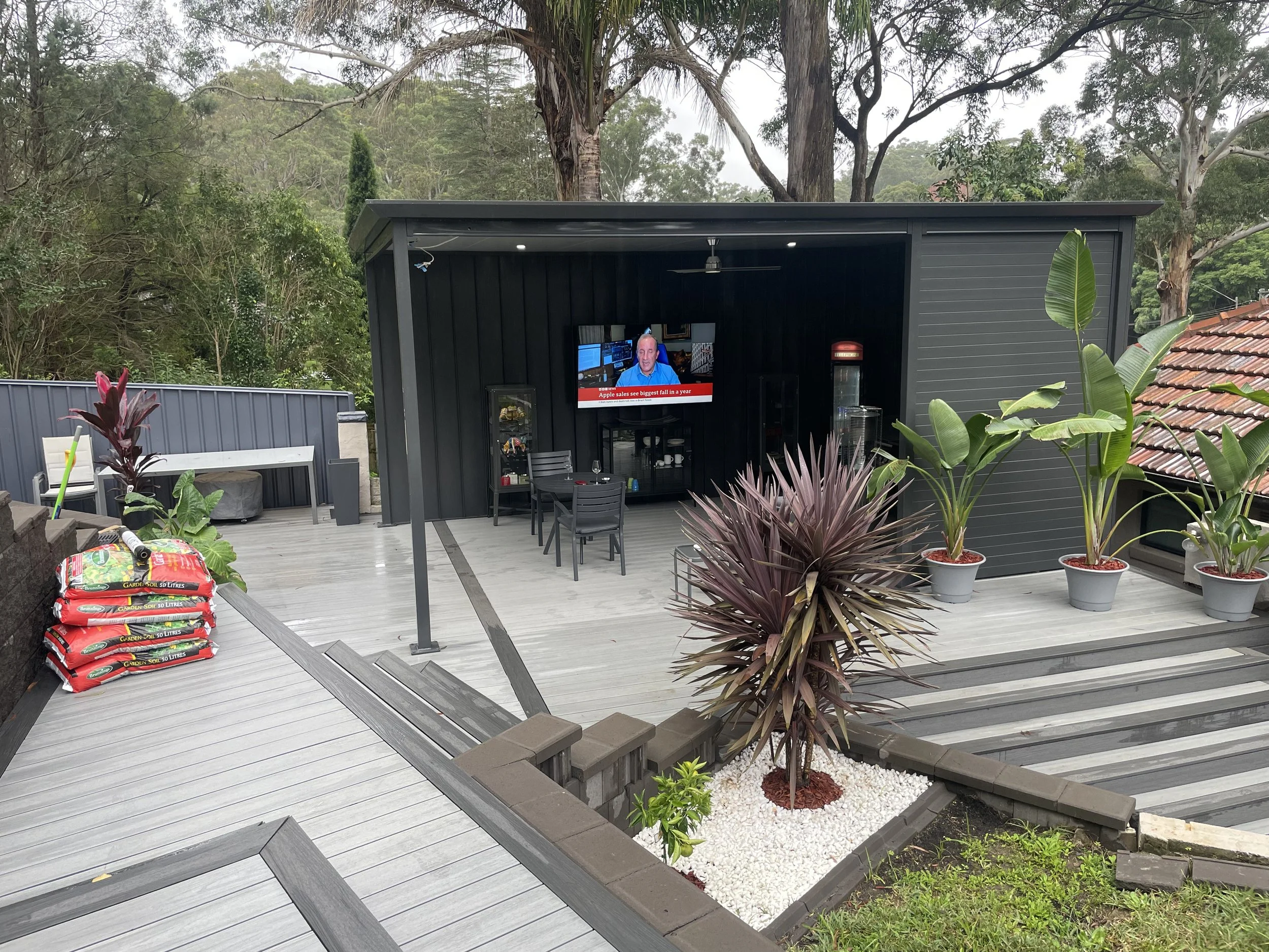 A backyard patio area with wpc decking, a black outdoor structure with a TV, and various potted plants including banana plants with large green leaves. Garden soil bags are stacked on the left side of the image.