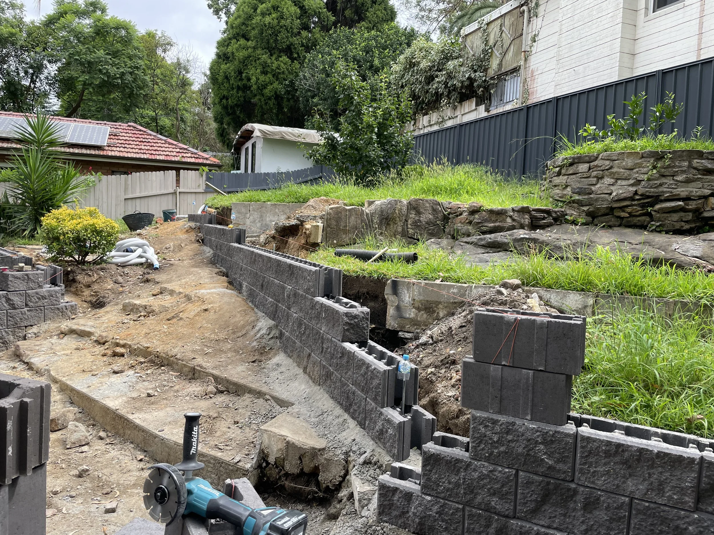 A backyard under construction with ongoing retaining wall construction using concrete blocks, construction tools, soil, and landscaping with trees and bushes.