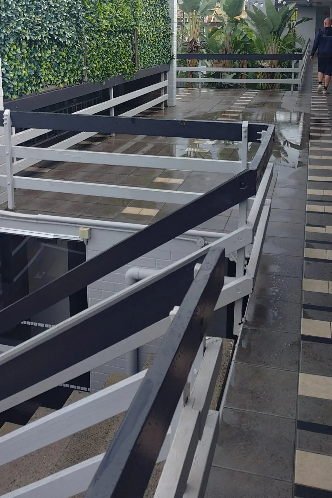 Empty outdoor balcony with old wood railings, wet tiled floor, and a man walking away in casual clothing.