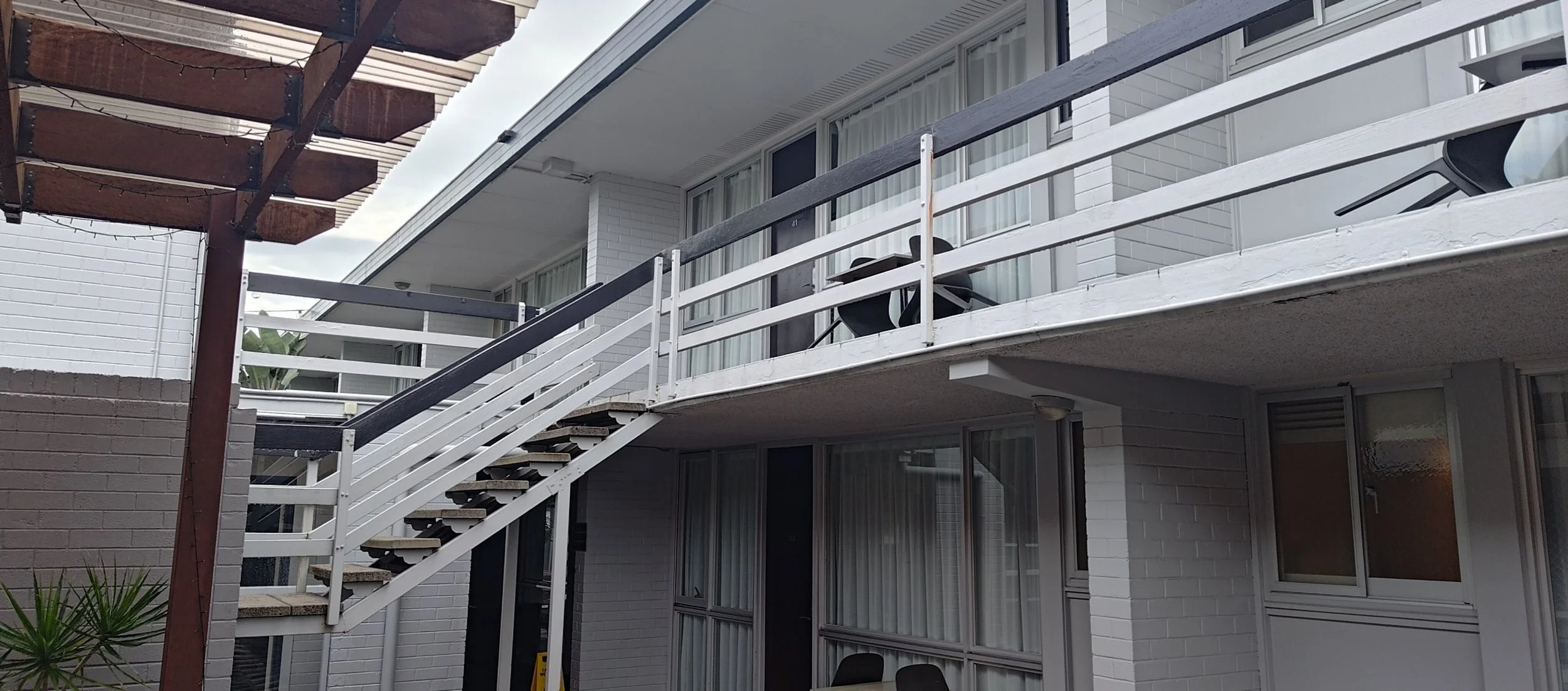 Hotel balcony with old white railing, black chairs, and a partially visible patio umbrella, part of a white brick building with large windows.