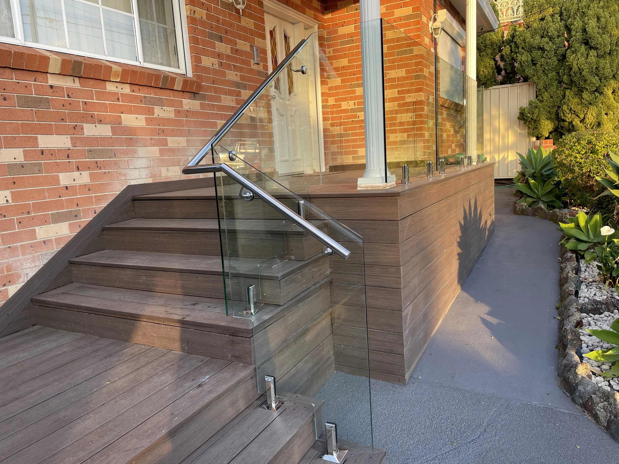 Wooden outdoor stairs with glass railing leading to a door of a brick house, surrounded by green plants, with a concrete pathway to the right.