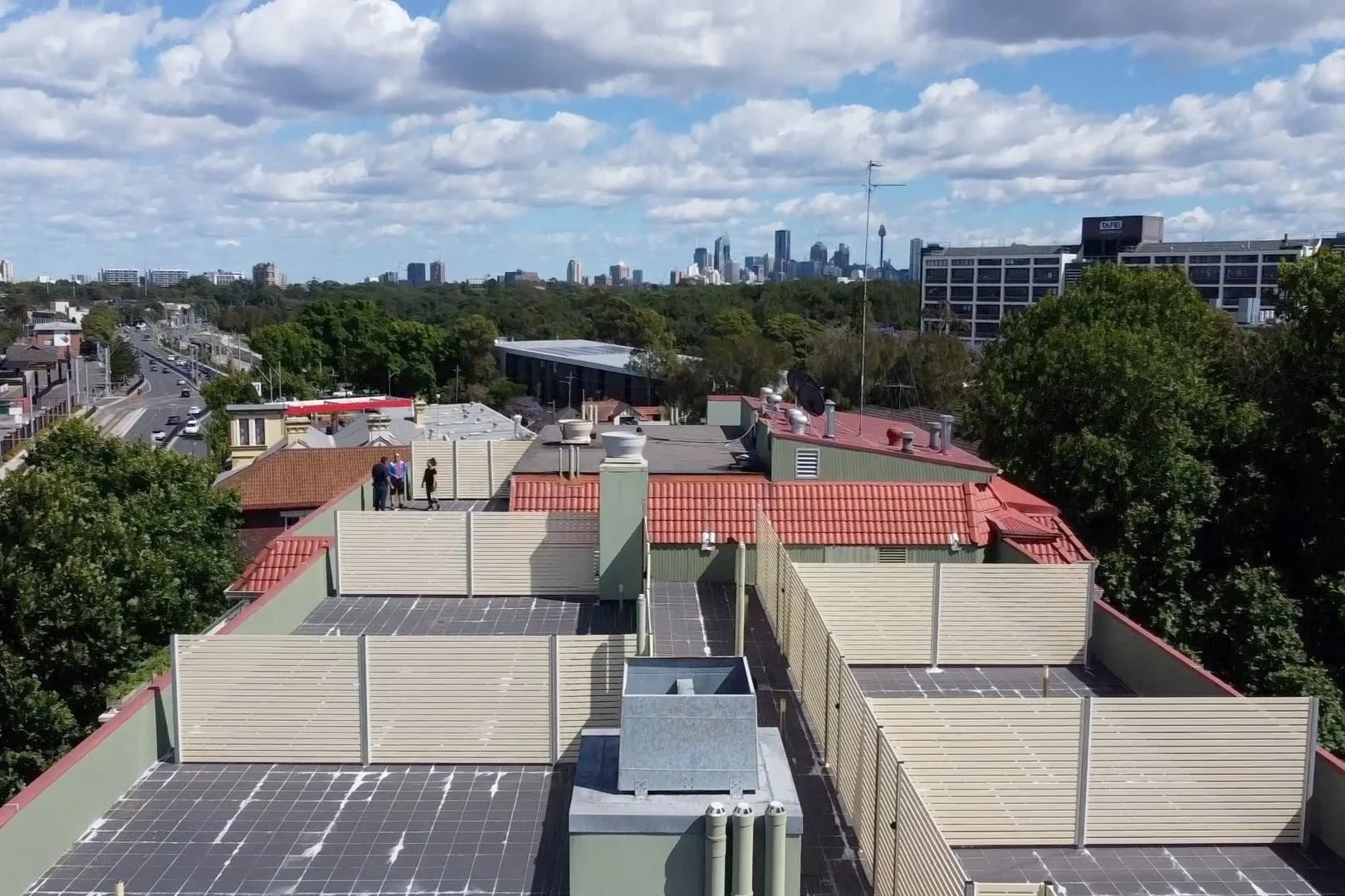 View from a rooftop showing solar panels, ventilation systems, and a few people standing on the roof. In the background, a city skyline with tall buildings, trees, and a partly cloudy sky.