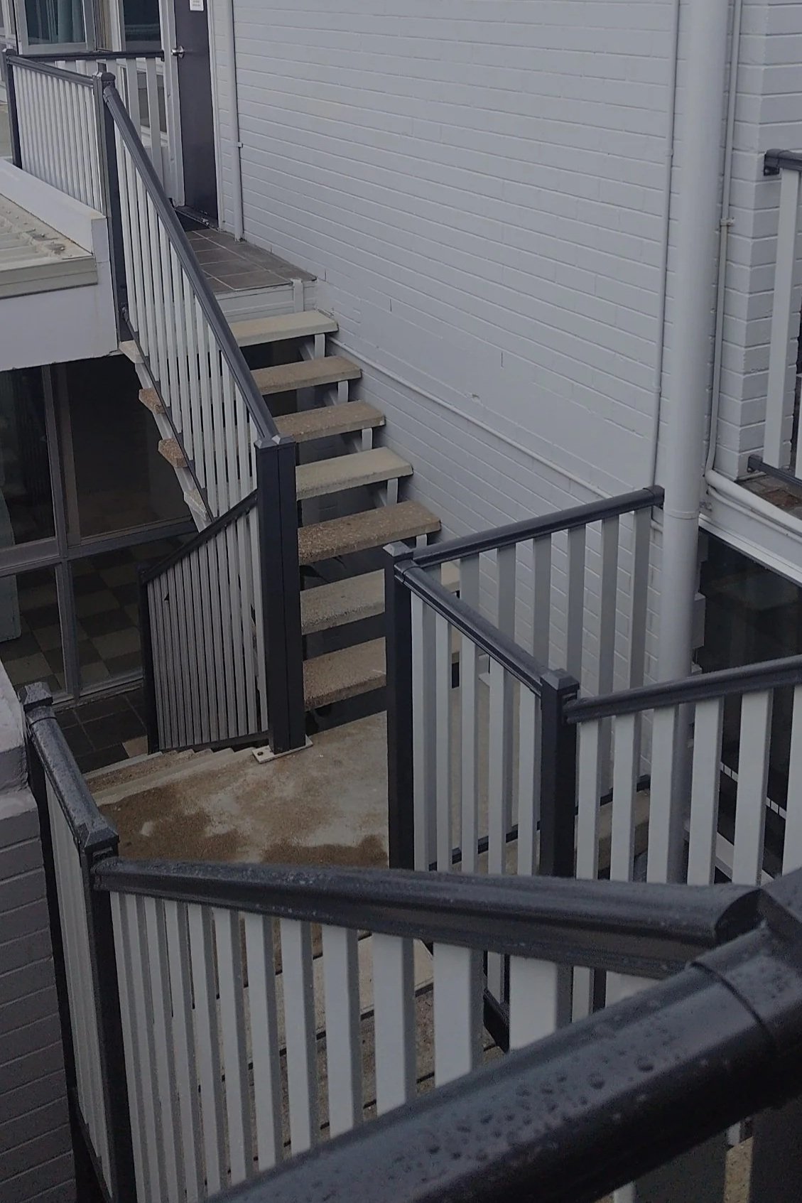 Outdoor view of a staircase with beige steps and a gray wpc railing, leading up to a door on the second floor of a white brick building.