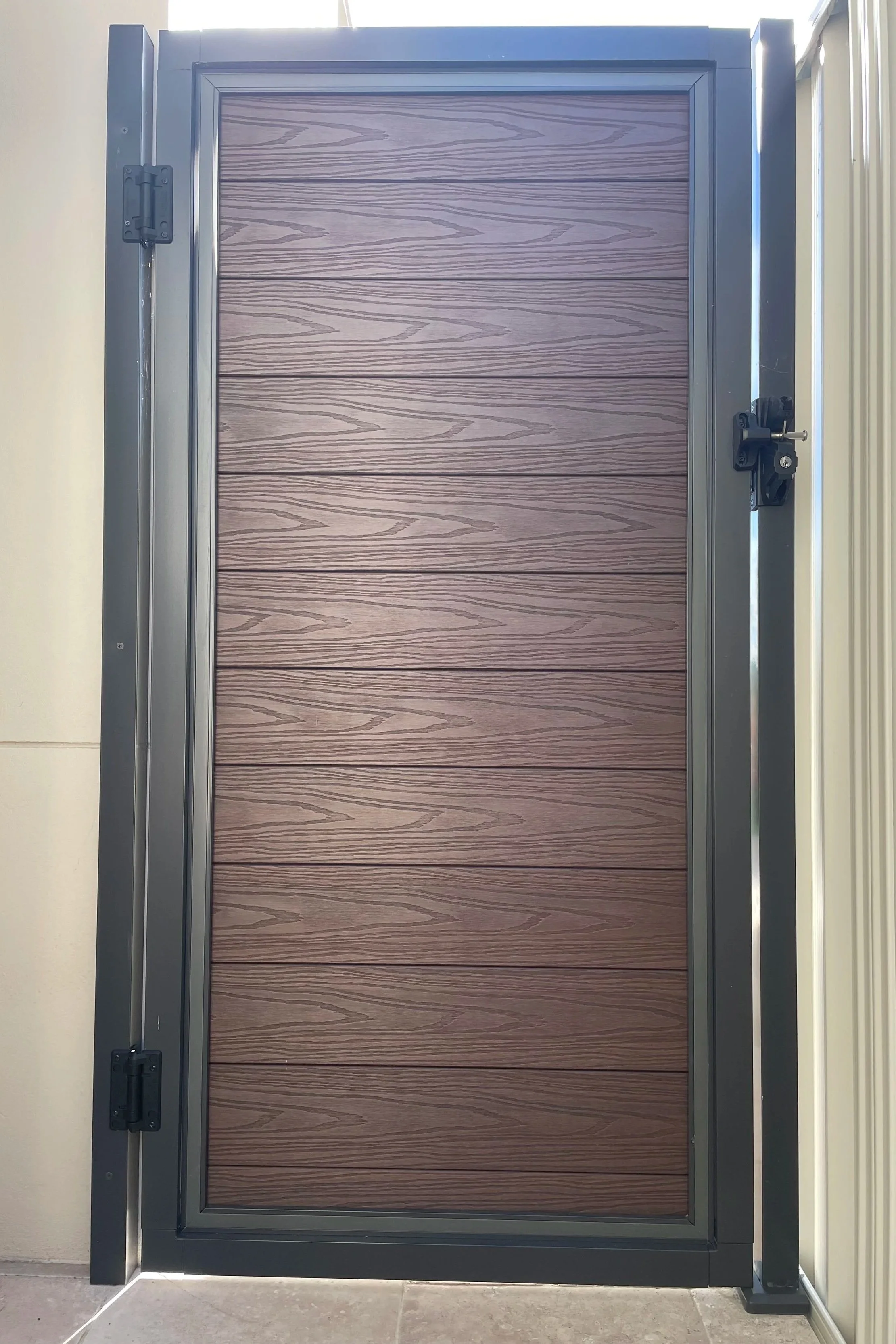 A large wood-plastic composite door with a dark aluminium frame and black hinges, installed in an interior setting with a light-colored wall and tiled floor.
