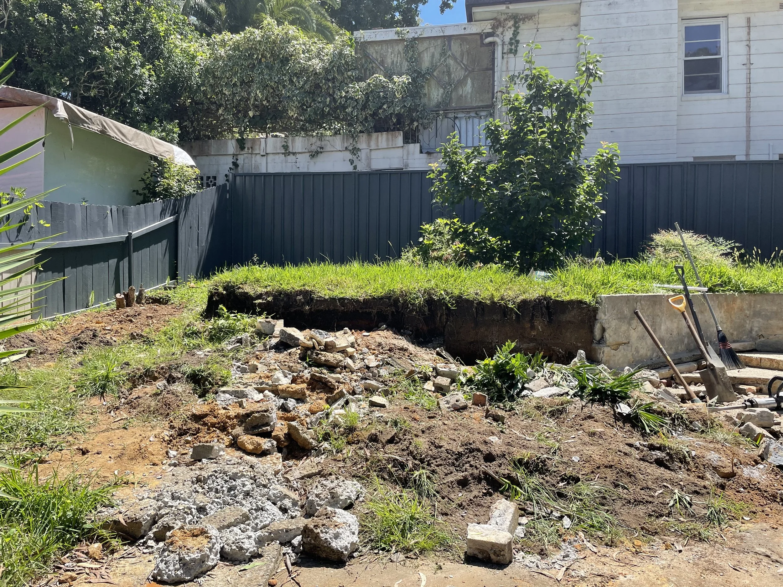 A backyard with green grass, a blue fence, and a small tree, with an excavated dirt patch and scattered rocks and construction tools in the foreground, and a white house and lush greenery in the background.