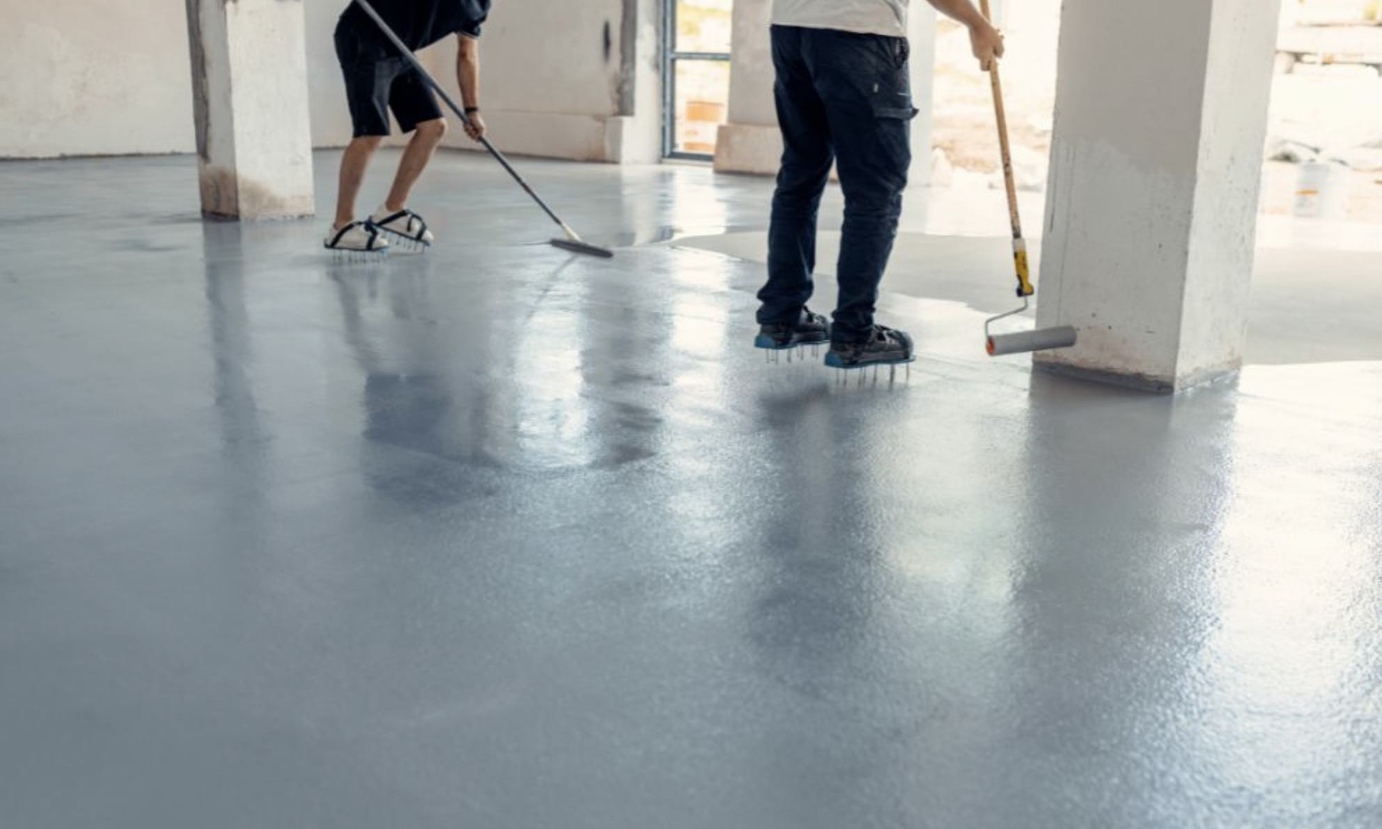 Two workers applying a fresh coat of gray epoxy to a concrete floor in a large, open indoor space with columns and a window.