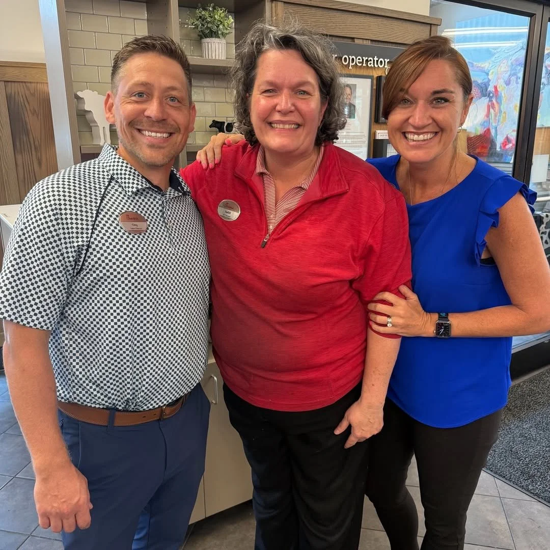Three happy people smiling and posing for a photo indoors, with two women and one man. The man is on the left, wearing a checked shirt and brown belt. The woman in the middle is wearing red, and the woman on the right is wearing a bright blue top. They are standing close together, and the woman on the right has her arm around the woman in the middle.