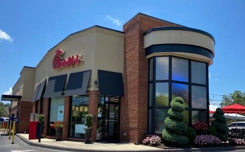 Chick-fil-A restaurant with brick and beige exterior, large blue-tinted windows, and decorative topiary bushes outside under a blue sky.