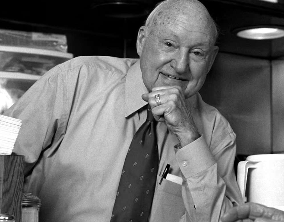 Black and white photo of an elderly man with a bald head, wearing a button-up shirt and a tie, smiling and resting his chin on his hand.