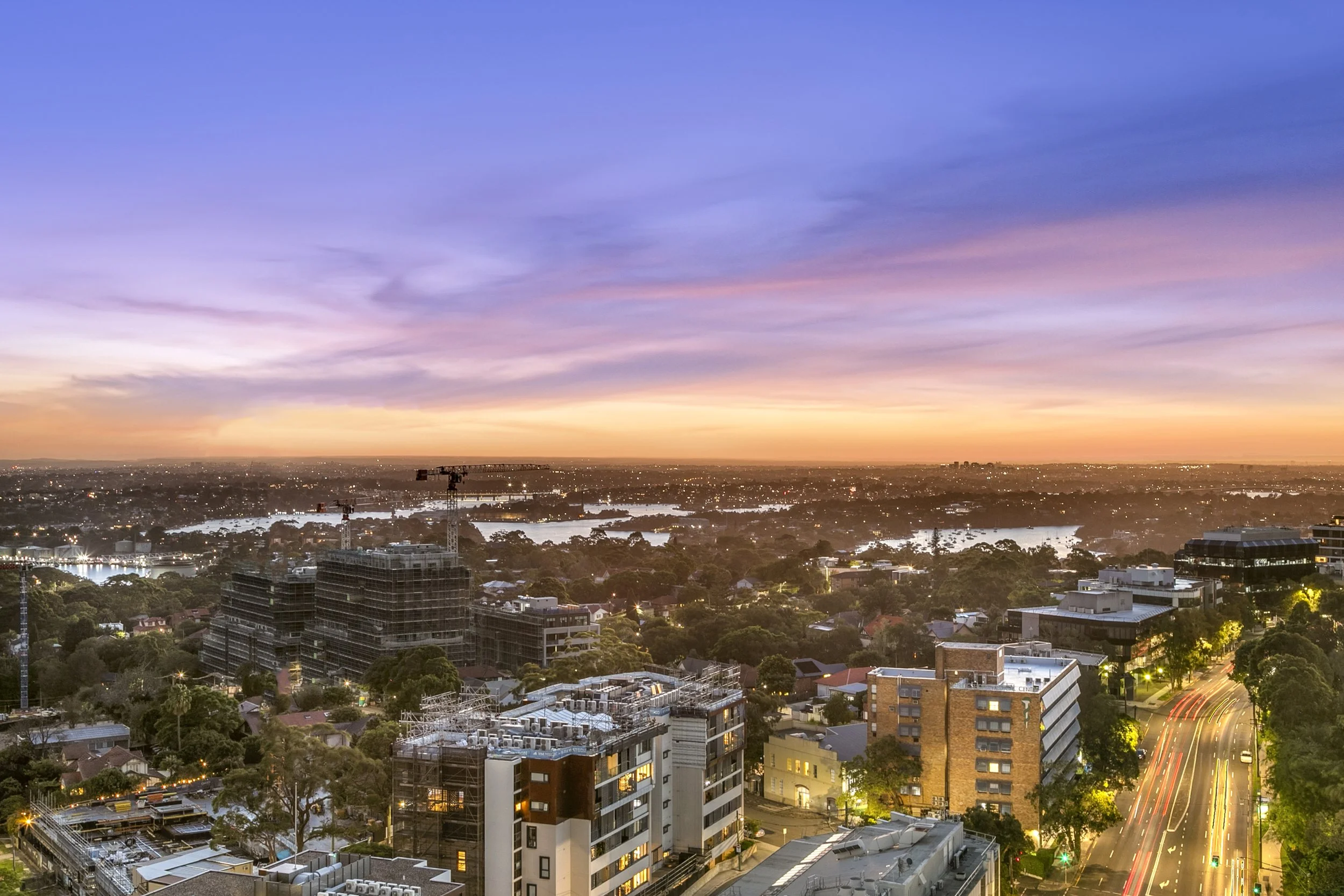 A cityscape at sunset showing buildings, streets, trees, and a purple-orange sky with clouds.