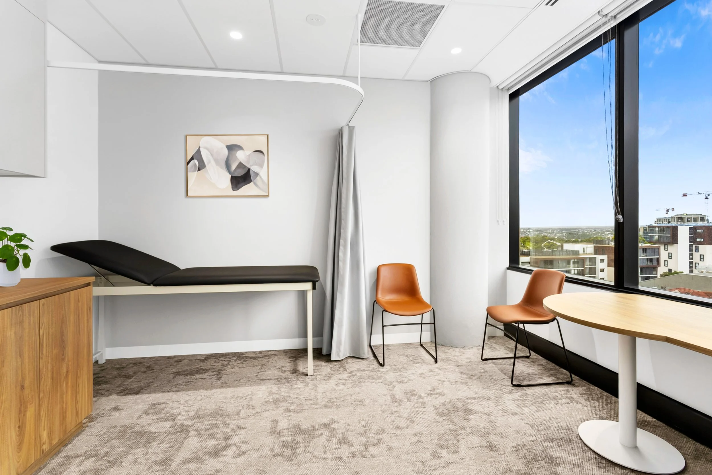 Medical examination room with a treatment bed, two orange chairs, a wooden table, a window with city view, and abstract wall art.