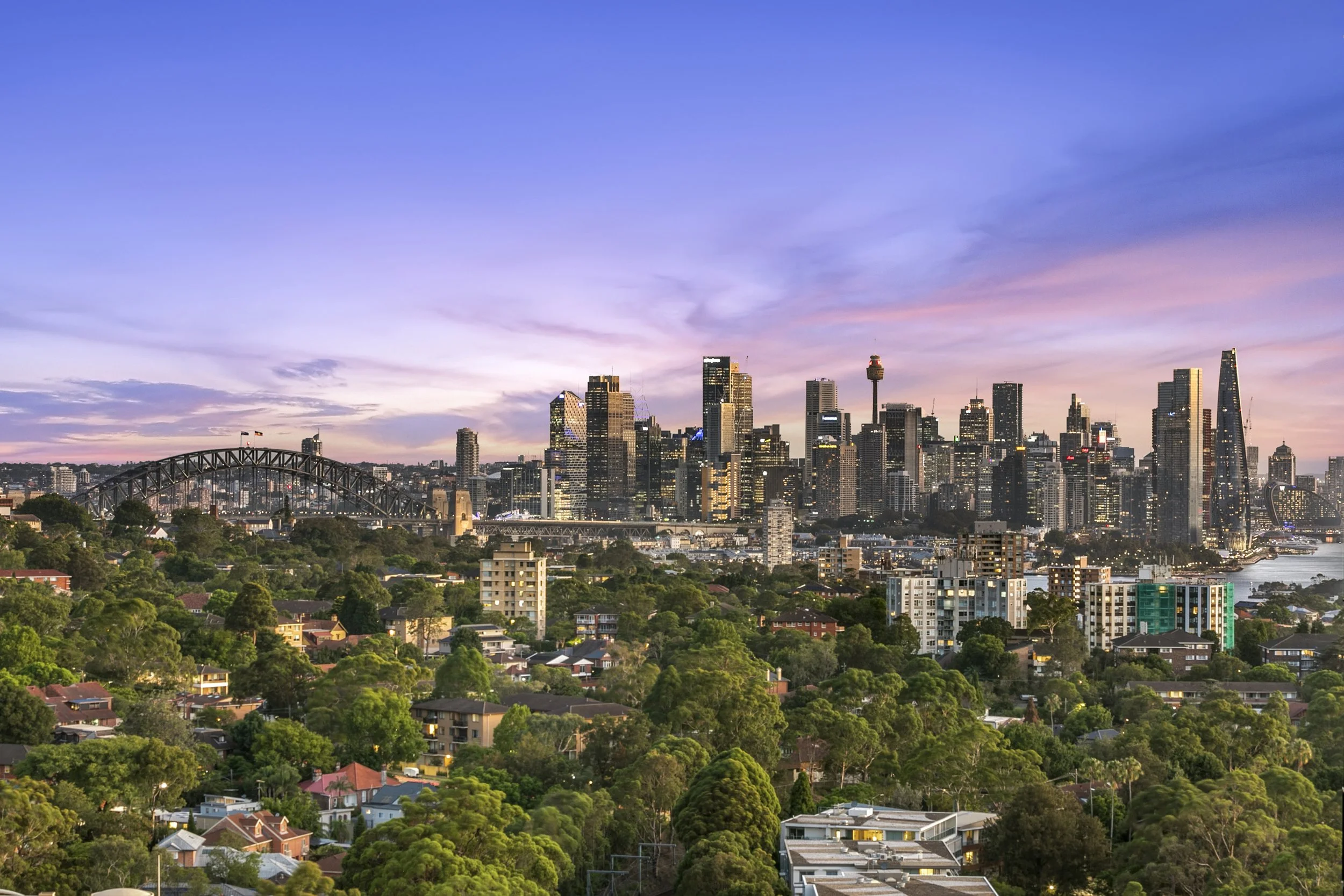 Sydney city skyline with tall skyscrapers and Sydney Harbour Bridge during a pink and purple sunset, with St Leonards residential and green areas in the foreground.