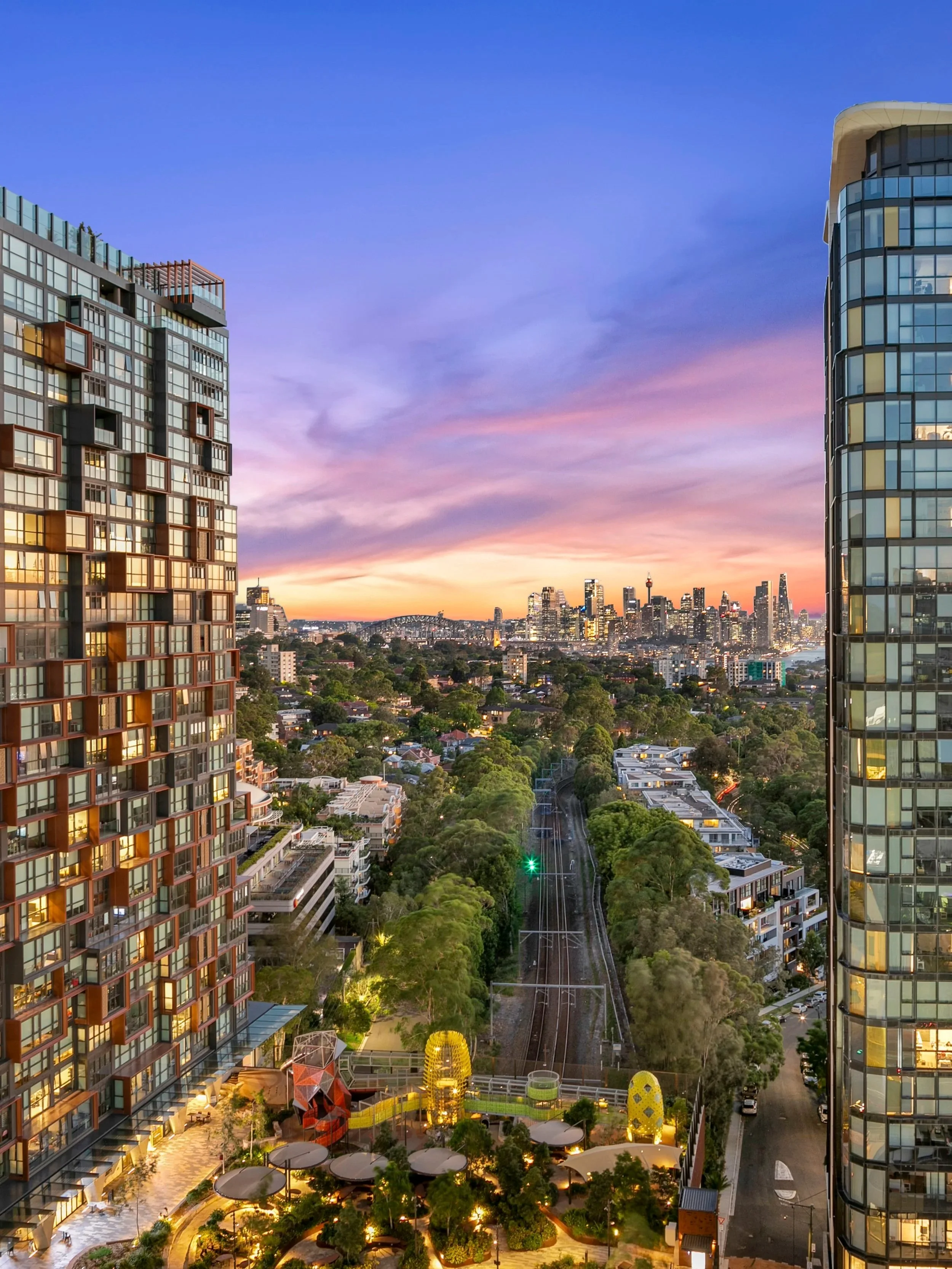St Leonards views with Sydney CBD skyline at sunset with modern high-rise buildings, a park with trees, and a railway track in the foreground.