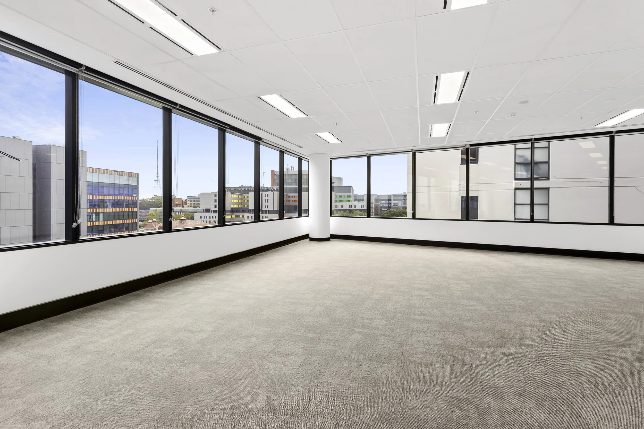 Empty office space with large windows, beige carpet, white walls, and ceiling lights, with views outside.