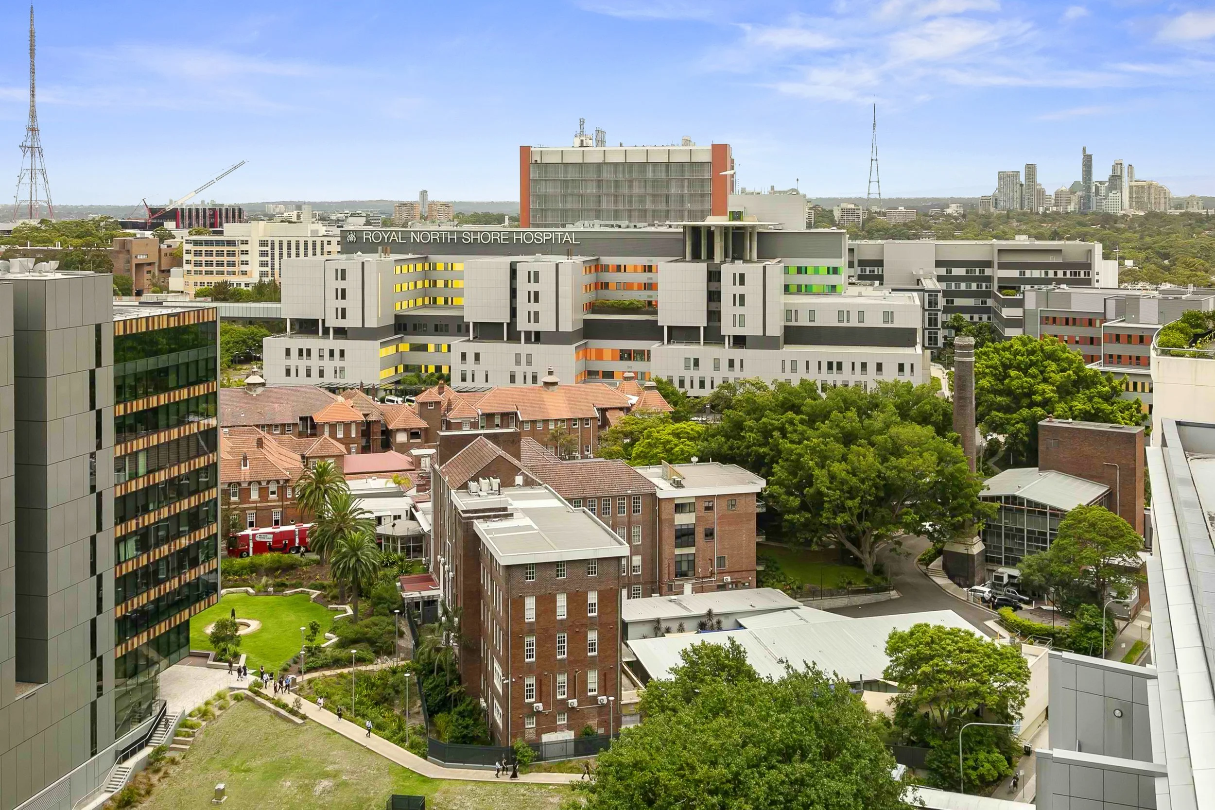 A view with a modern hospital building labeled 'Royal North Shore Hospital' in the background, surrounded by trees, brick buildings, and other modern structures, under a partly cloudy sky.