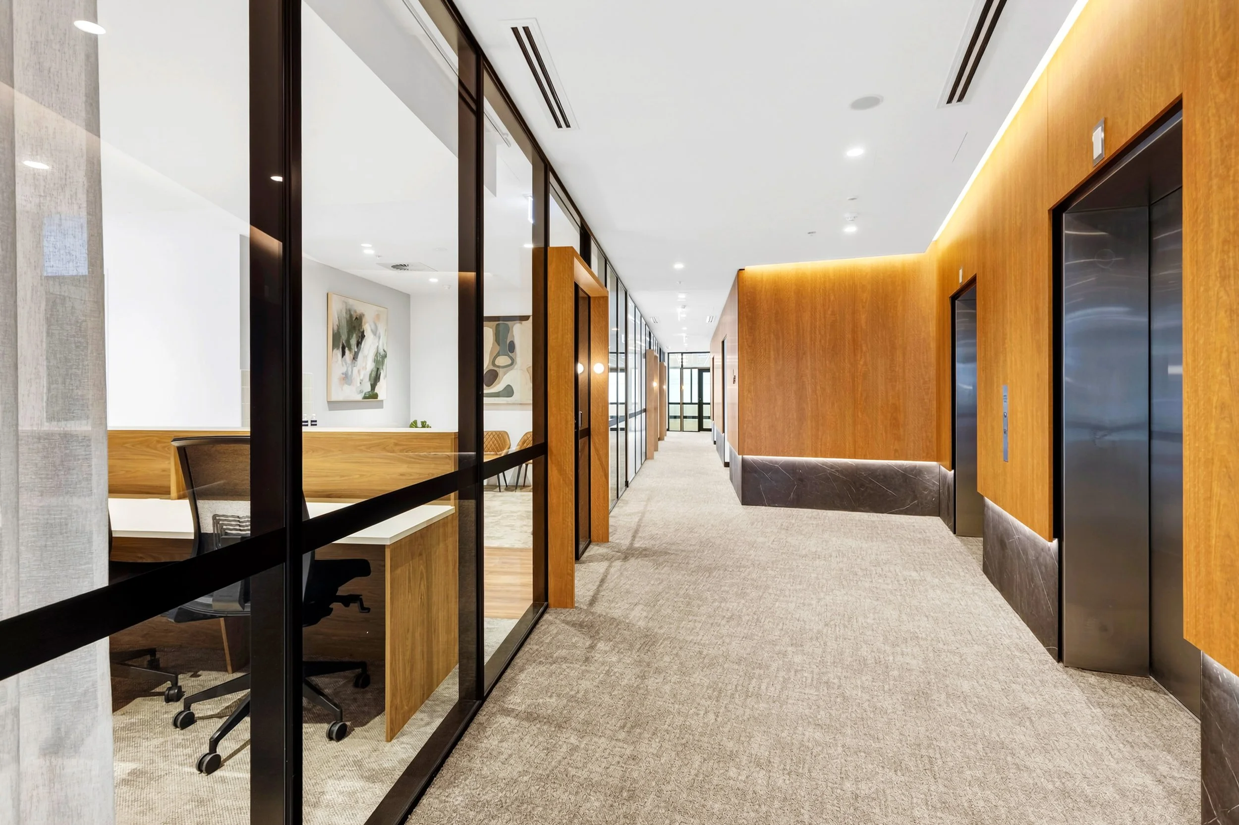 Modern office hallway with glass-walls on the left, wooden and marble accents, and elevator doors on the right, illuminated with overhead lighting.