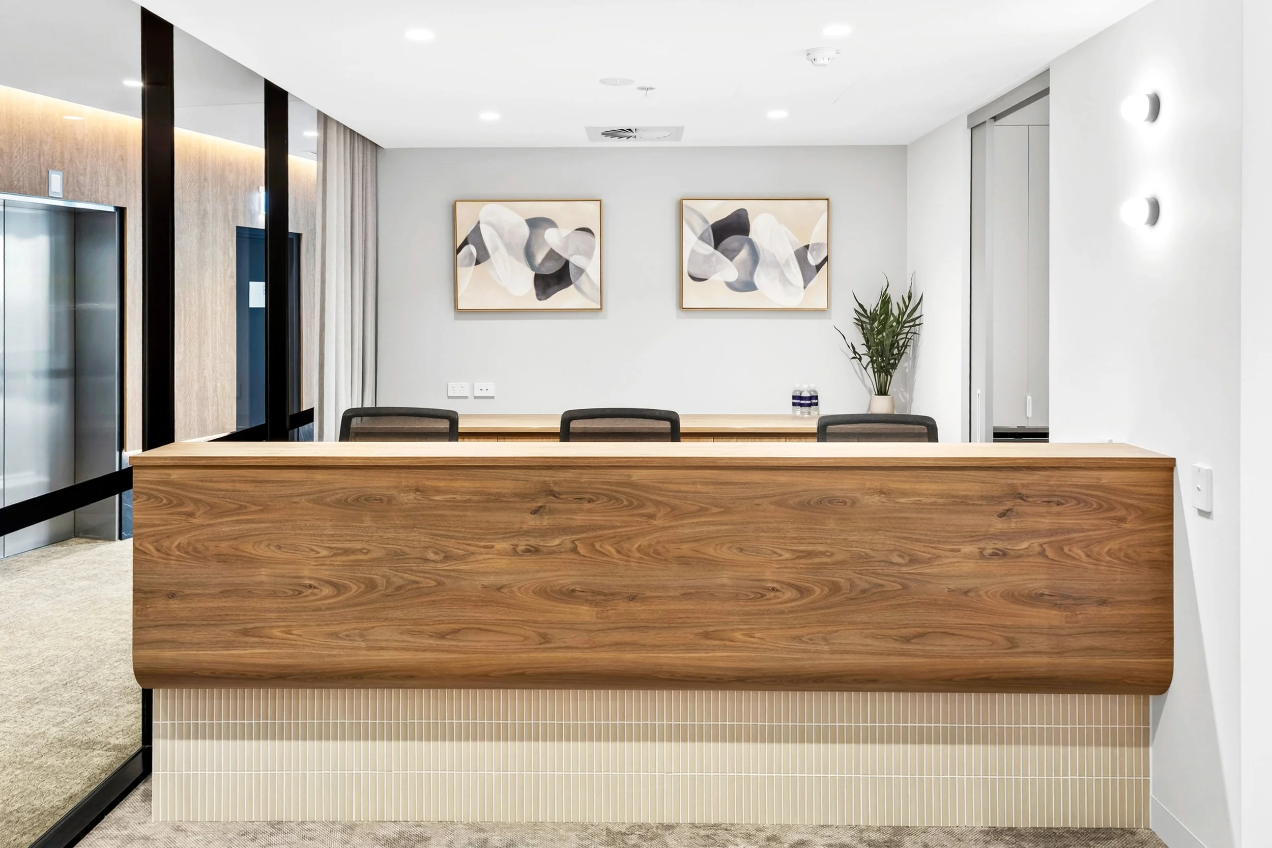 Modern office reception area with a wooden desk, black chairs, abstract art on the white wall, and a tall potted plant.
